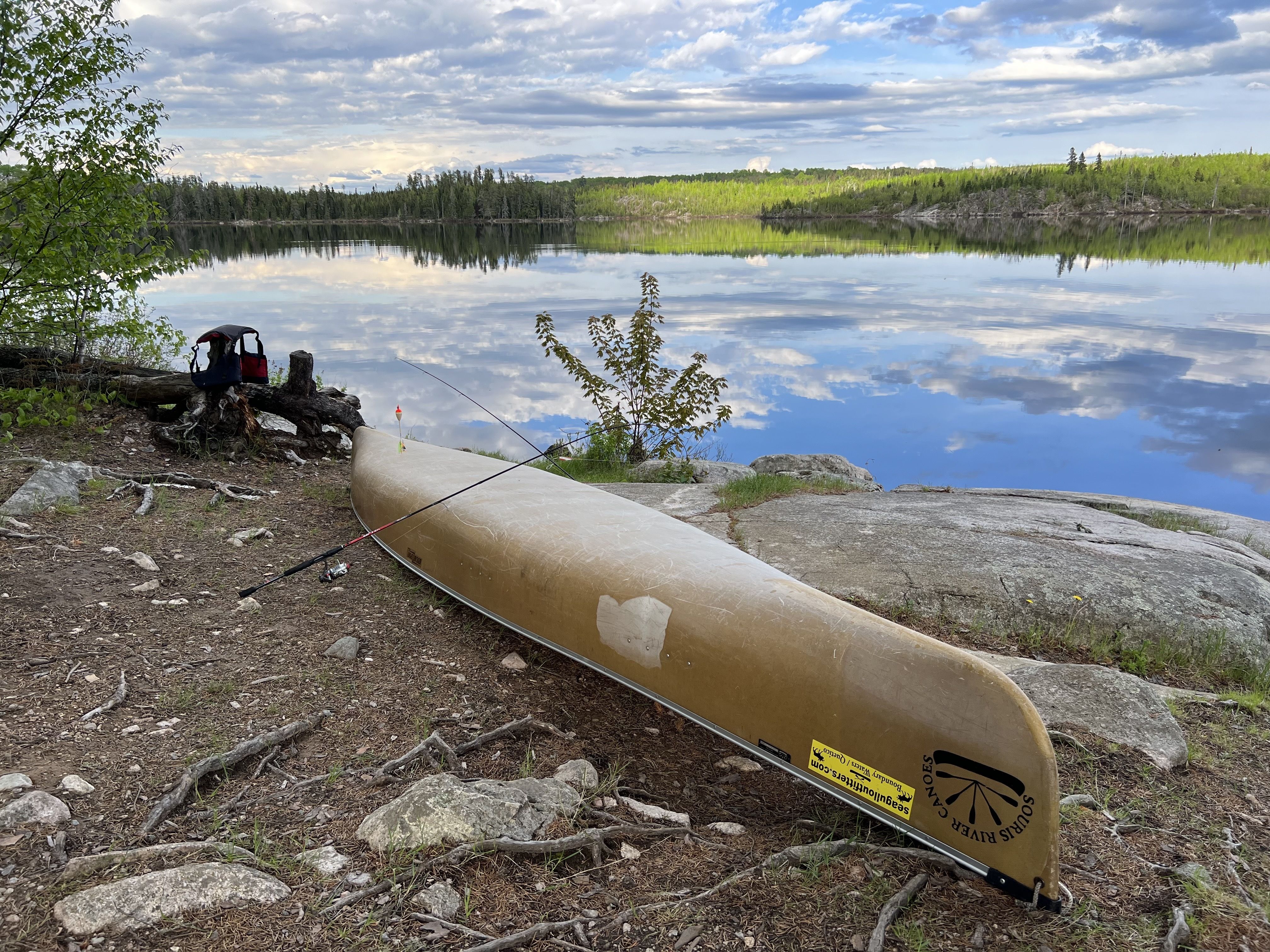 A brown kevlar canoe on the shore of a lake with fishing rods draped over the top of it