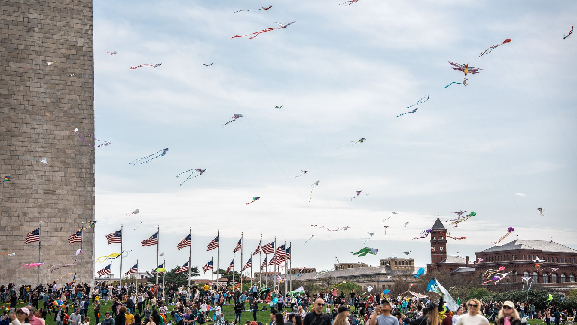Wide park scene with a crowd, colorful kites in the sky, rows of American flags on poles, a tall stone wall on the left, and brick buildings with a clock tower in the distance.