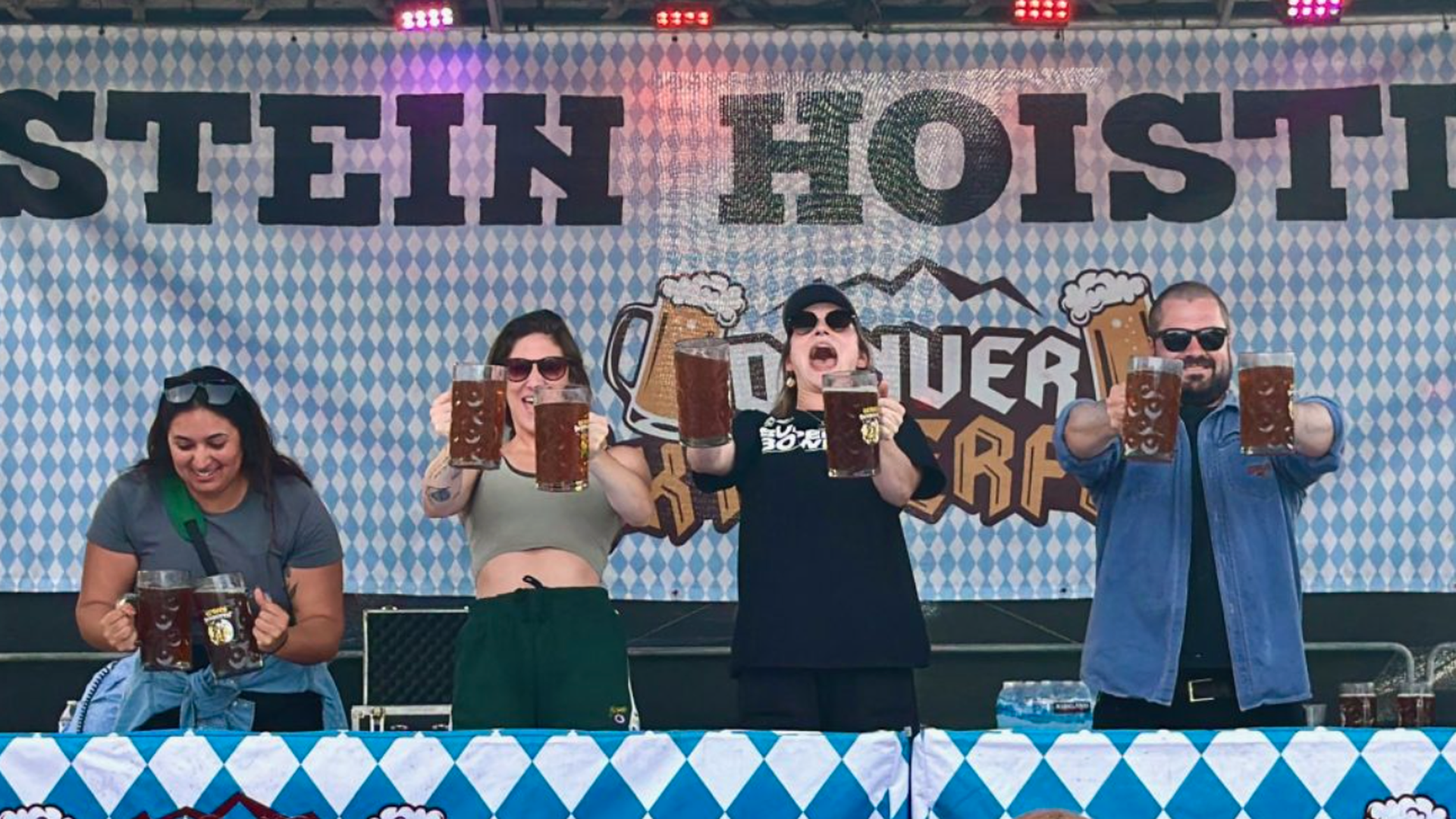 Four people on a stage with a blue-and-white diamond backdrop that says "STEIN HOIST", each raising large beer mugs in a celebratory stein-hoisting photo.
