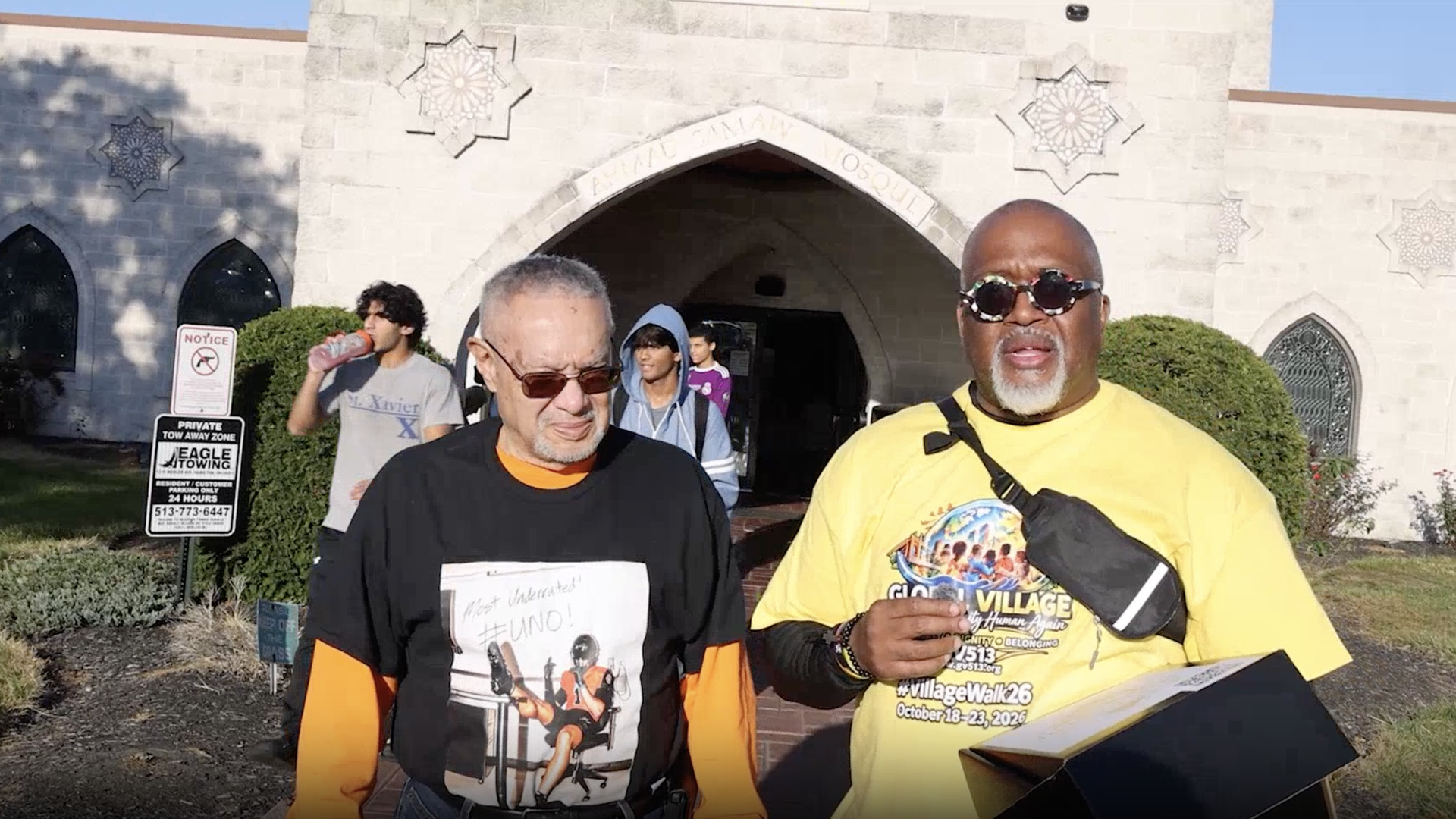 Two men stand outside a light gray stone building with an arched entrance and star motifs. One wears a yellow shirt and sunglasses; the other a black shirt with orange sleeves as people exit behind.