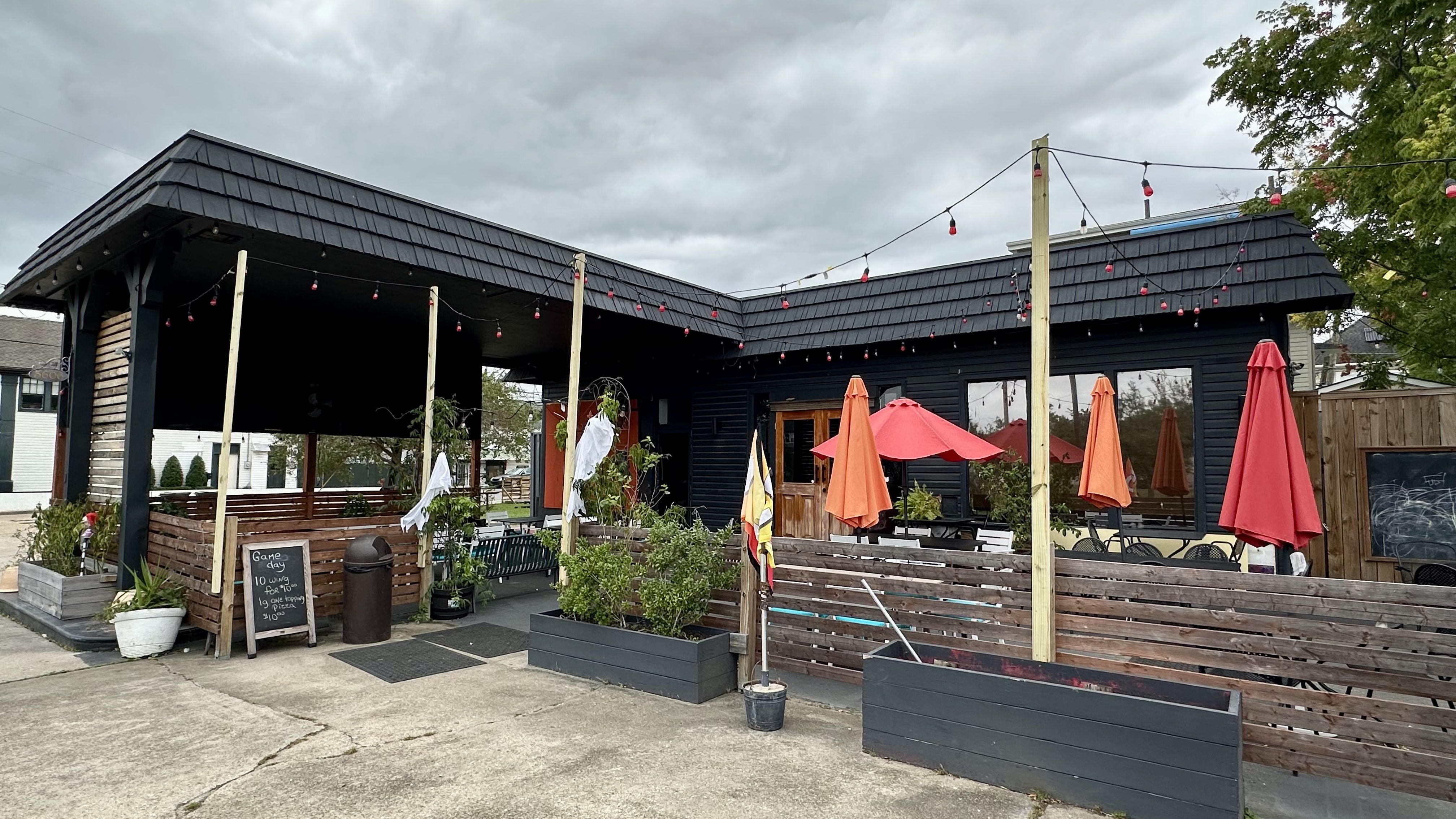 A black wooden cafe with a covered patio, string lights with red bulbs, and orange patio umbrellas. Outdoor tables along wooden fences, potted plants, under an overcast sky.
