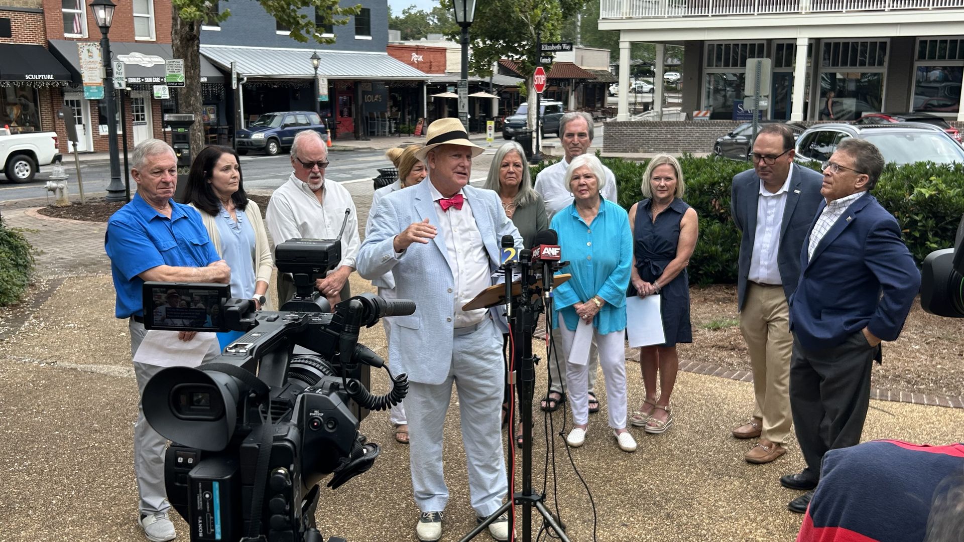 Man in light blue suit and straw hat with pink bow tie speaks at an outdoor press conference with microphones and camera, surrounded by a diverse group of people in casual and business attire.