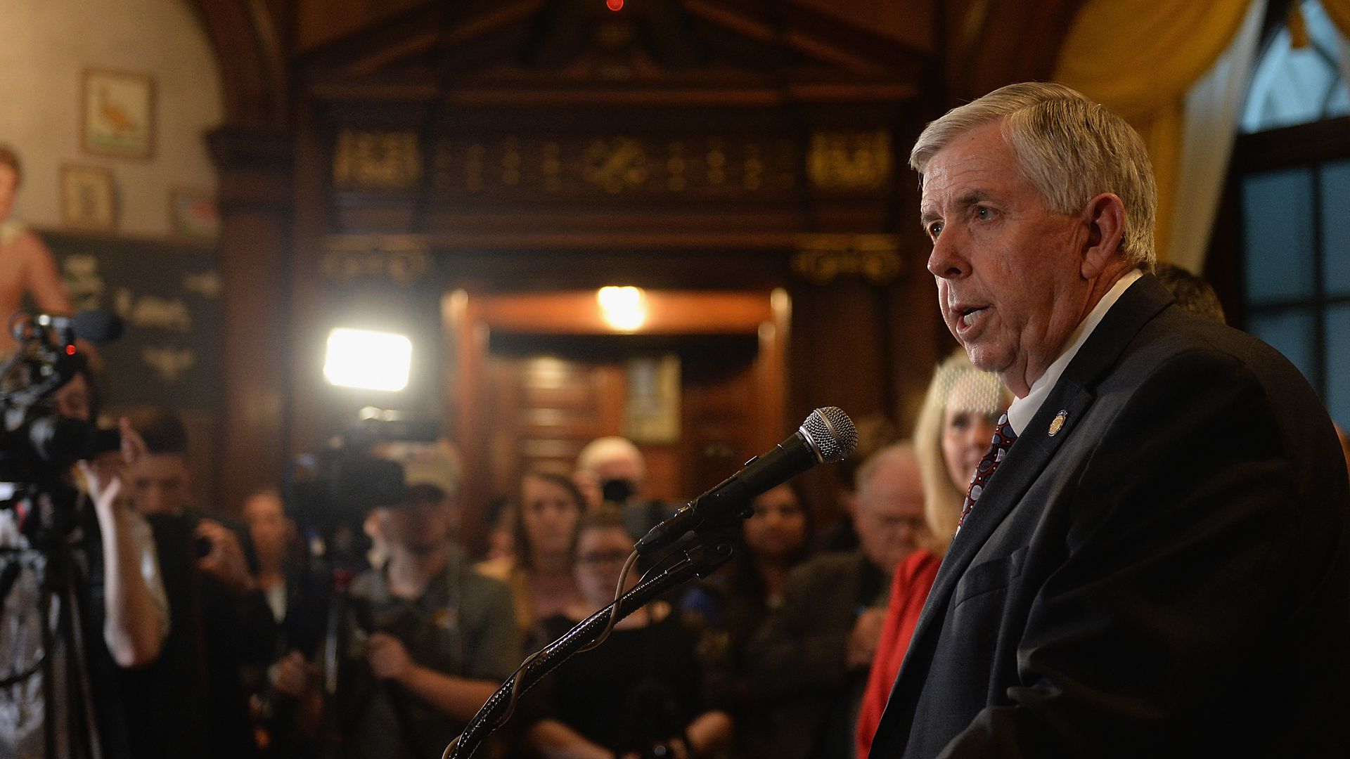 Photo of Mike Parson speaking from a podium to a room of media workers