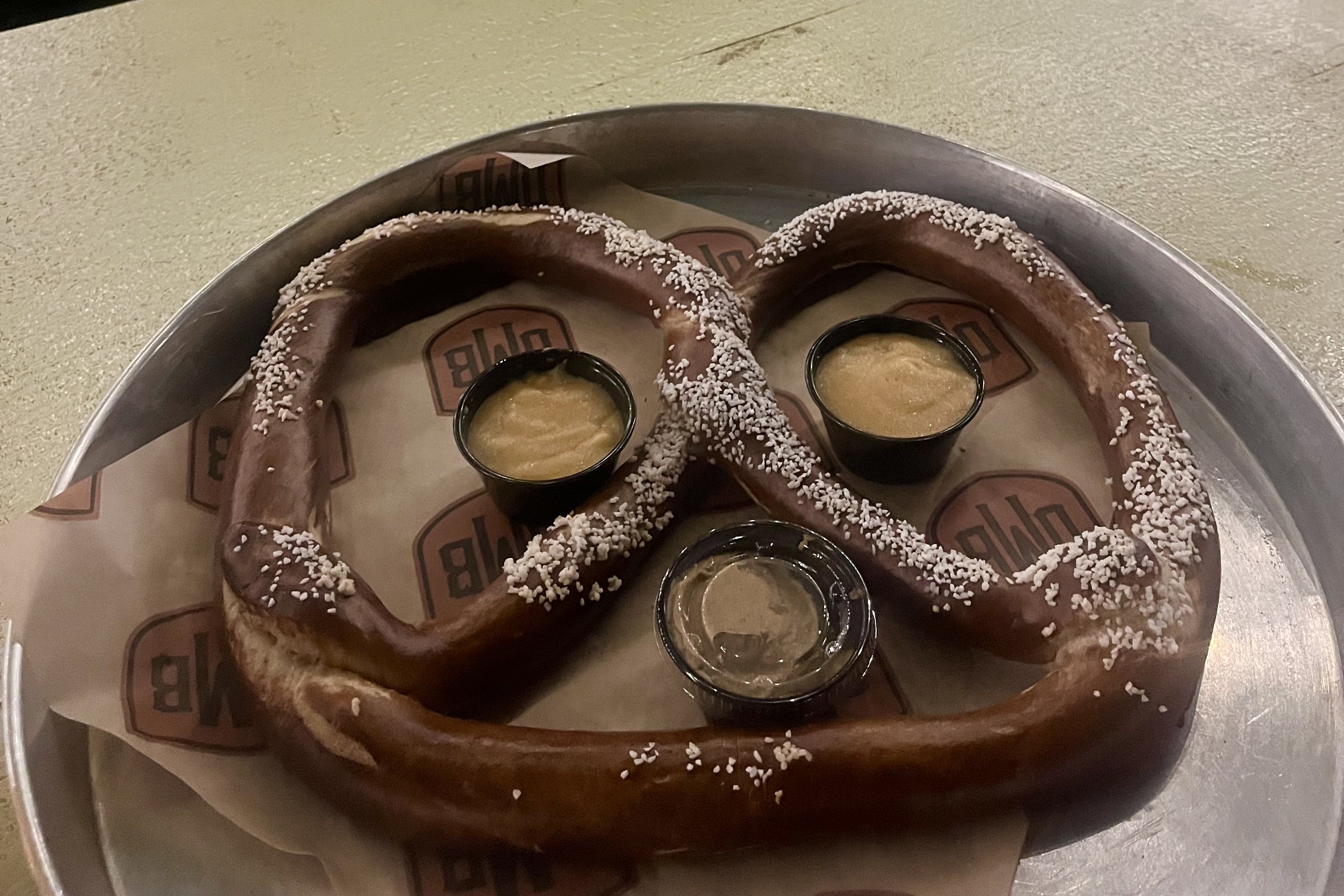 Two large dark brown pretzel rings with coarse white salt sit on a metal tray, surrounded by small black cups of yellow cheese dip; a branded paper liner is beneath.