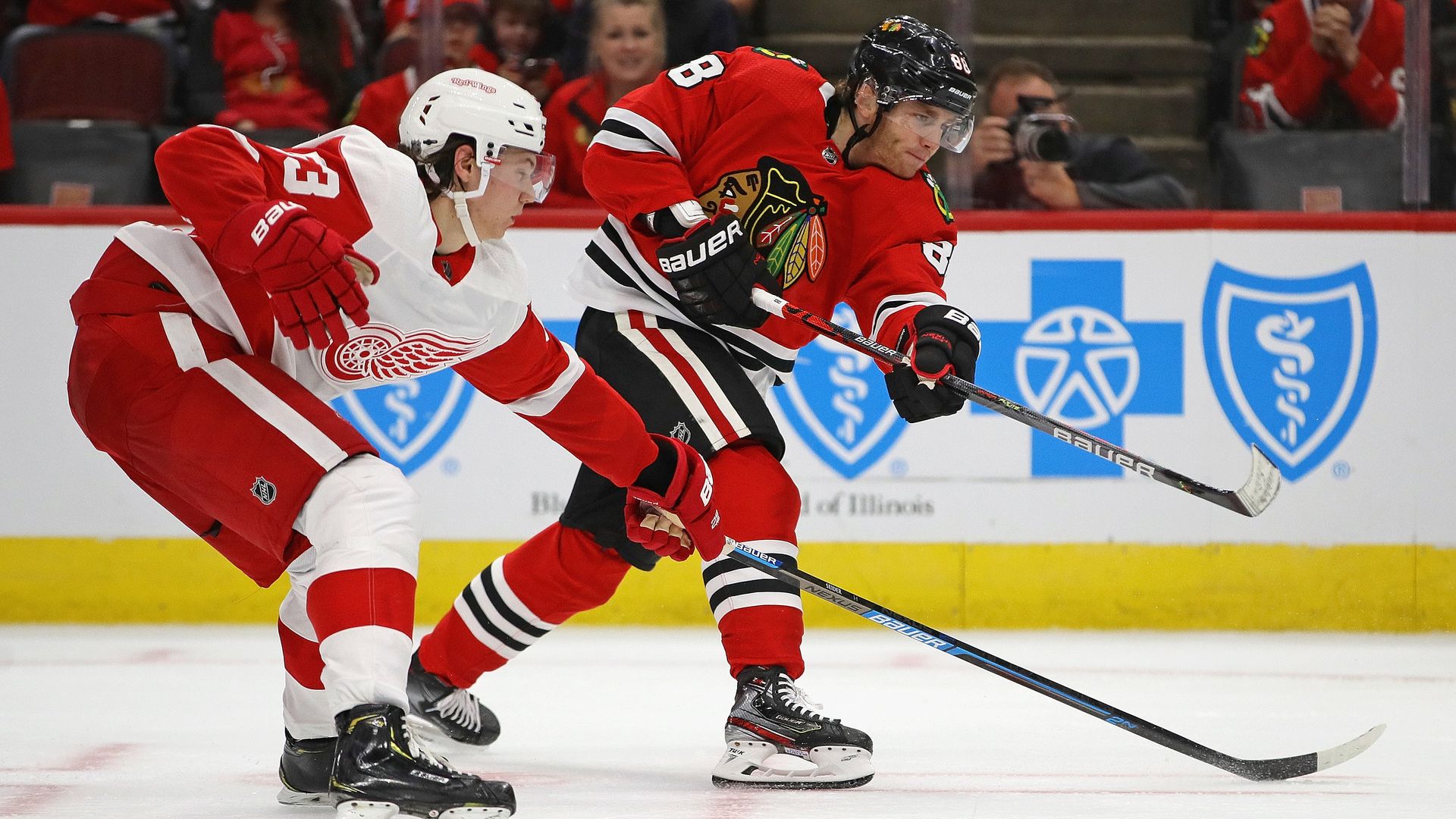 In this image, two hockey players face the right while skating across the ice, hockey sticks in front of them.