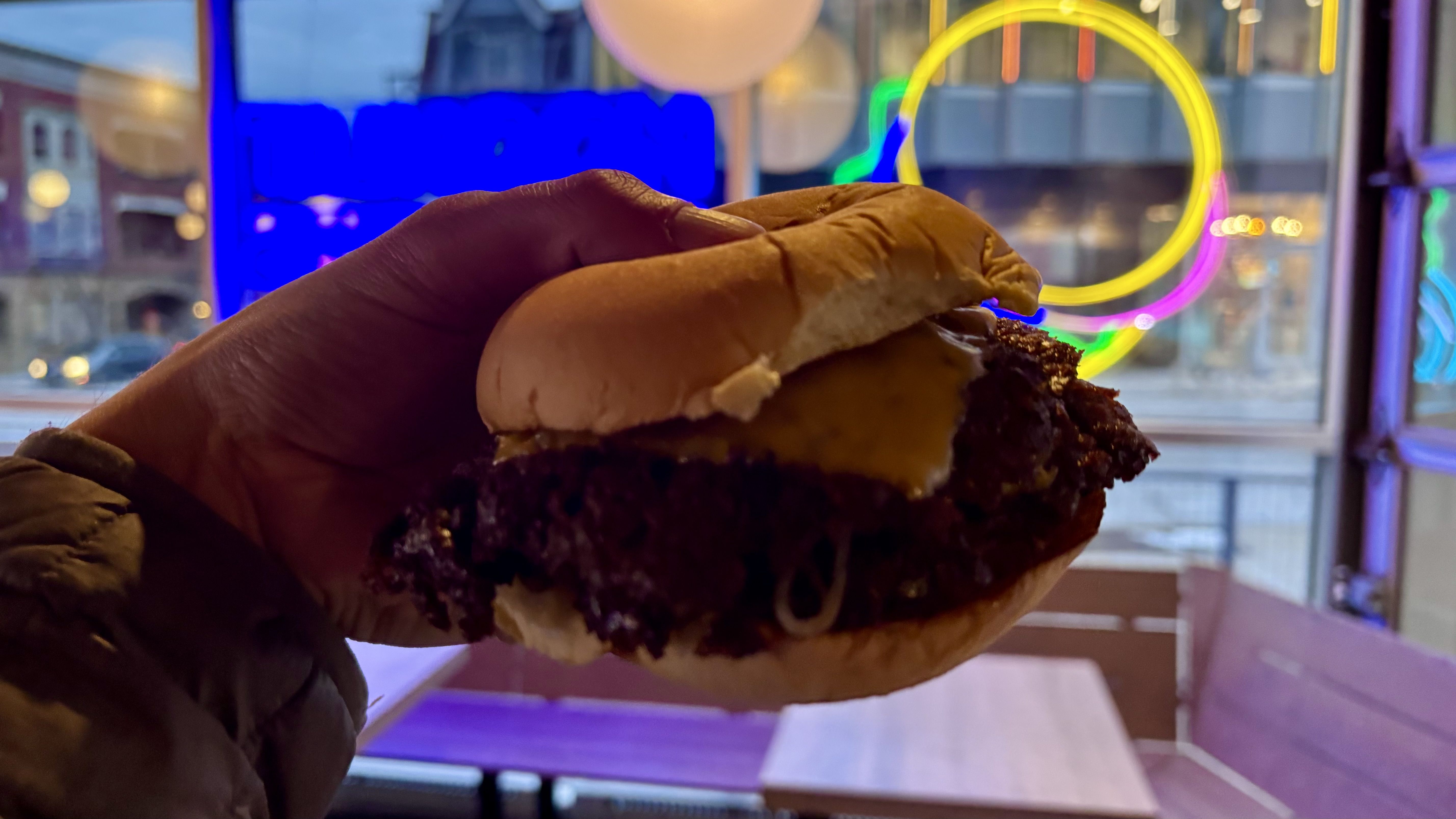 A hand holds a cheeseburger with melted cheese inside a dimly lit restaurant featuring neon lights in yellow, pink, blue, and green in the background during evening.