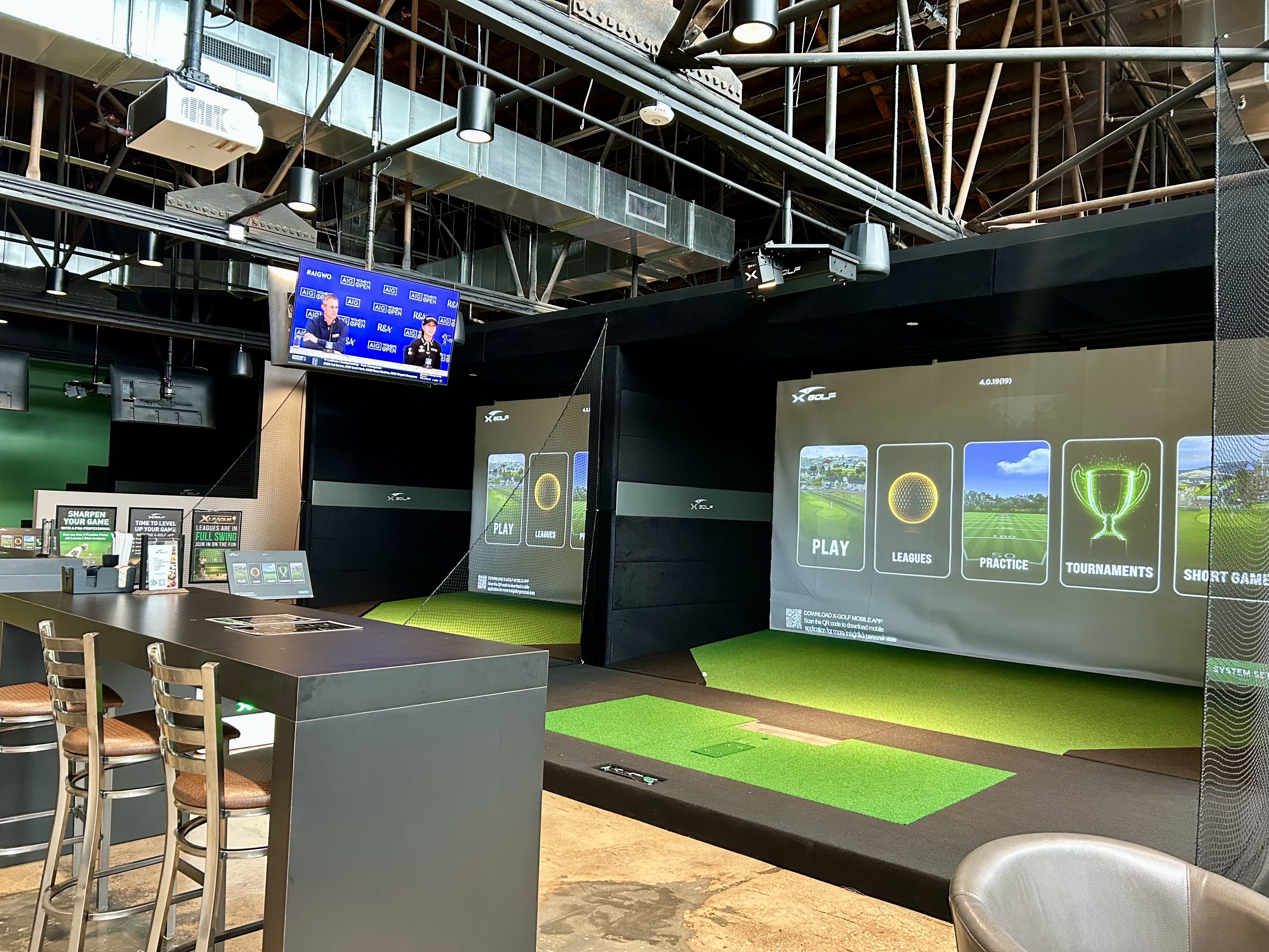 Indoor golf simulator setup with green artificial turf, two large projection screens showing golf menu options, stools, and a TV displaying a golf event in a modern facility.