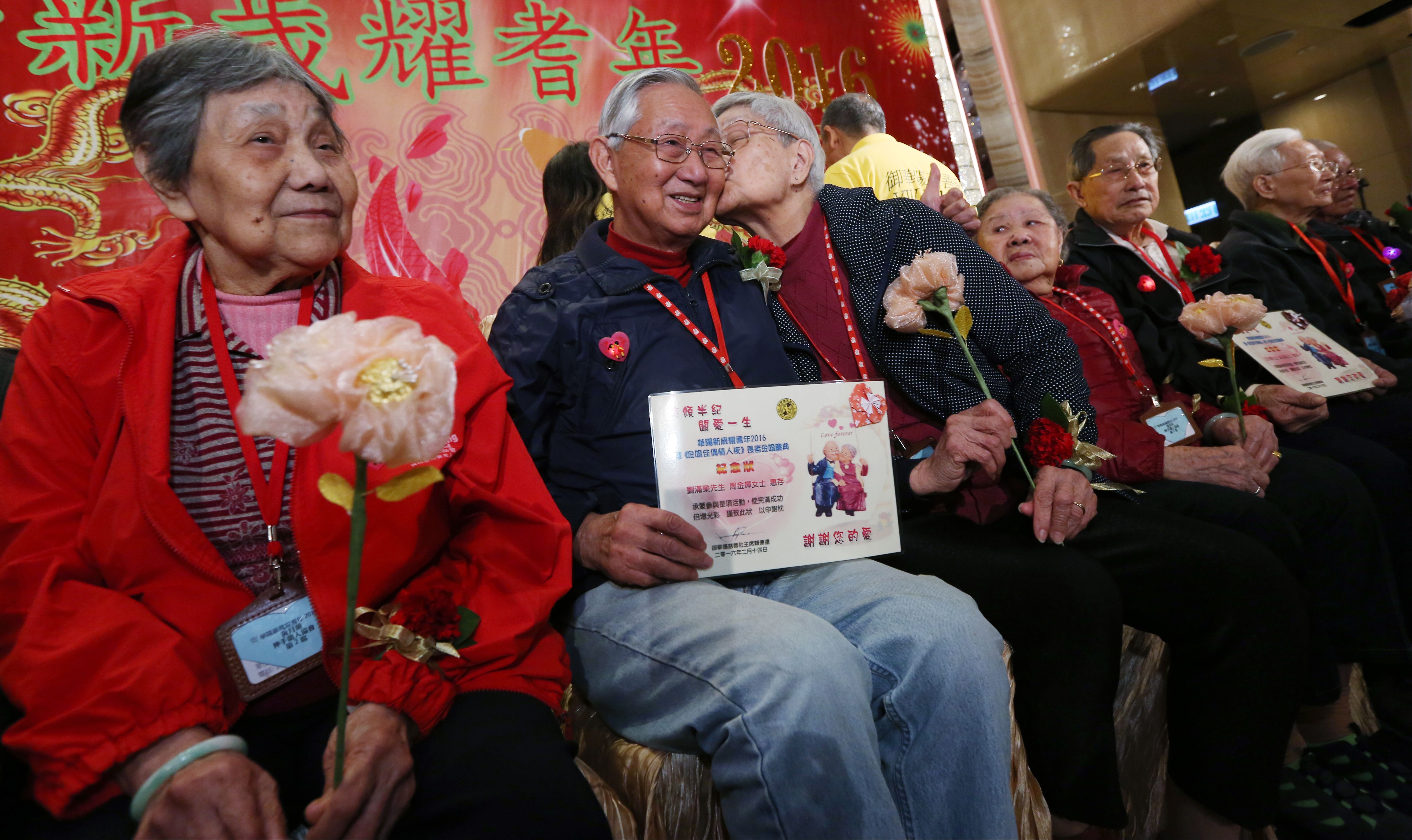 In this image, a line of elderly people sit with flowers and signs 