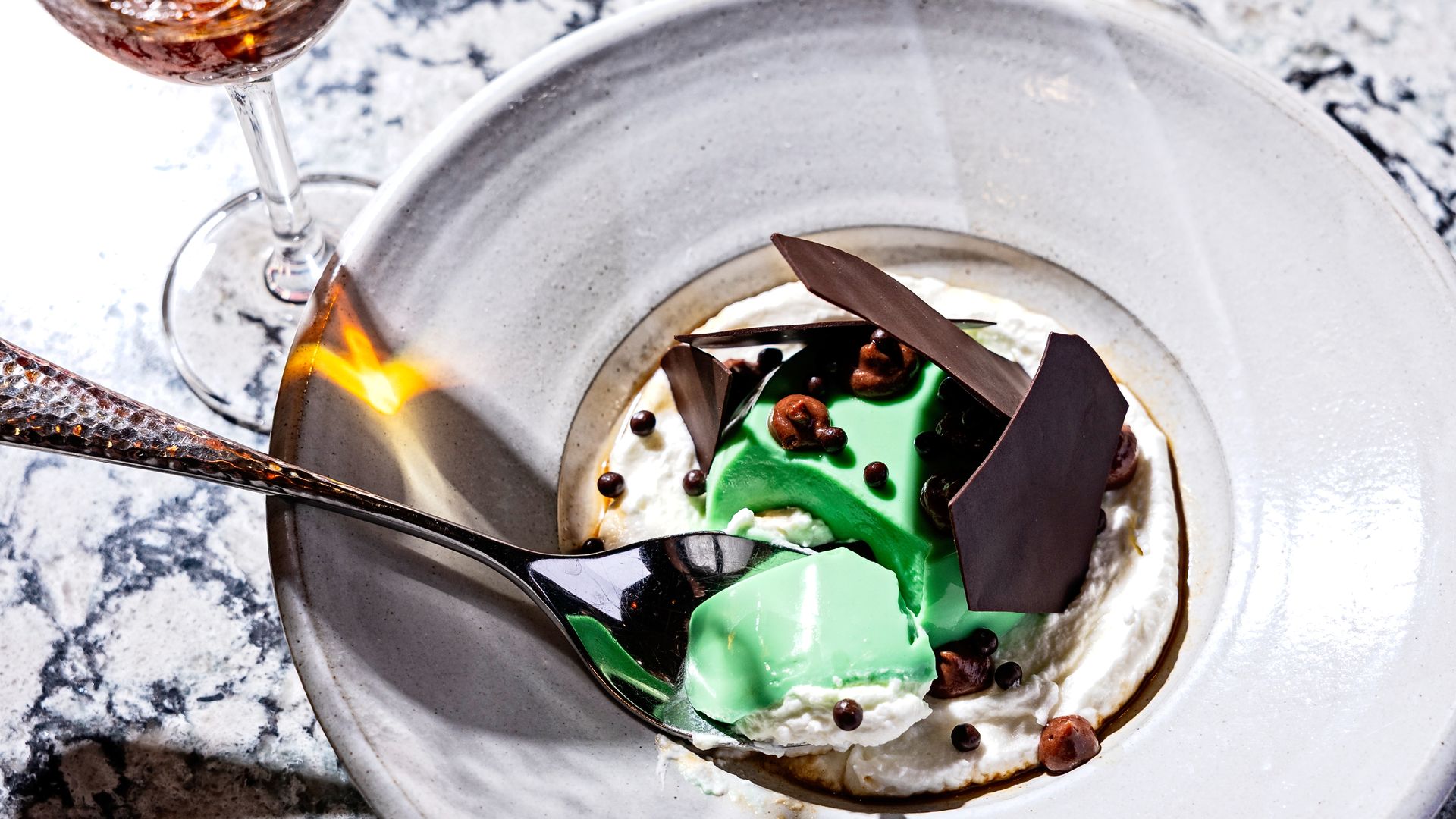 A green panna cotta in a white bowl with chocolate shavings next to a red cocktail 