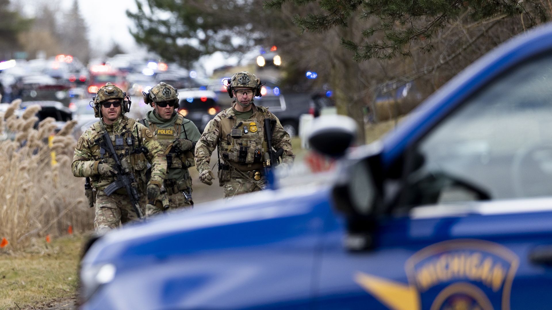 Law enforcement armed and wearing camo walk toward the camera. A Michigan police vehicle is pictured in the foreground