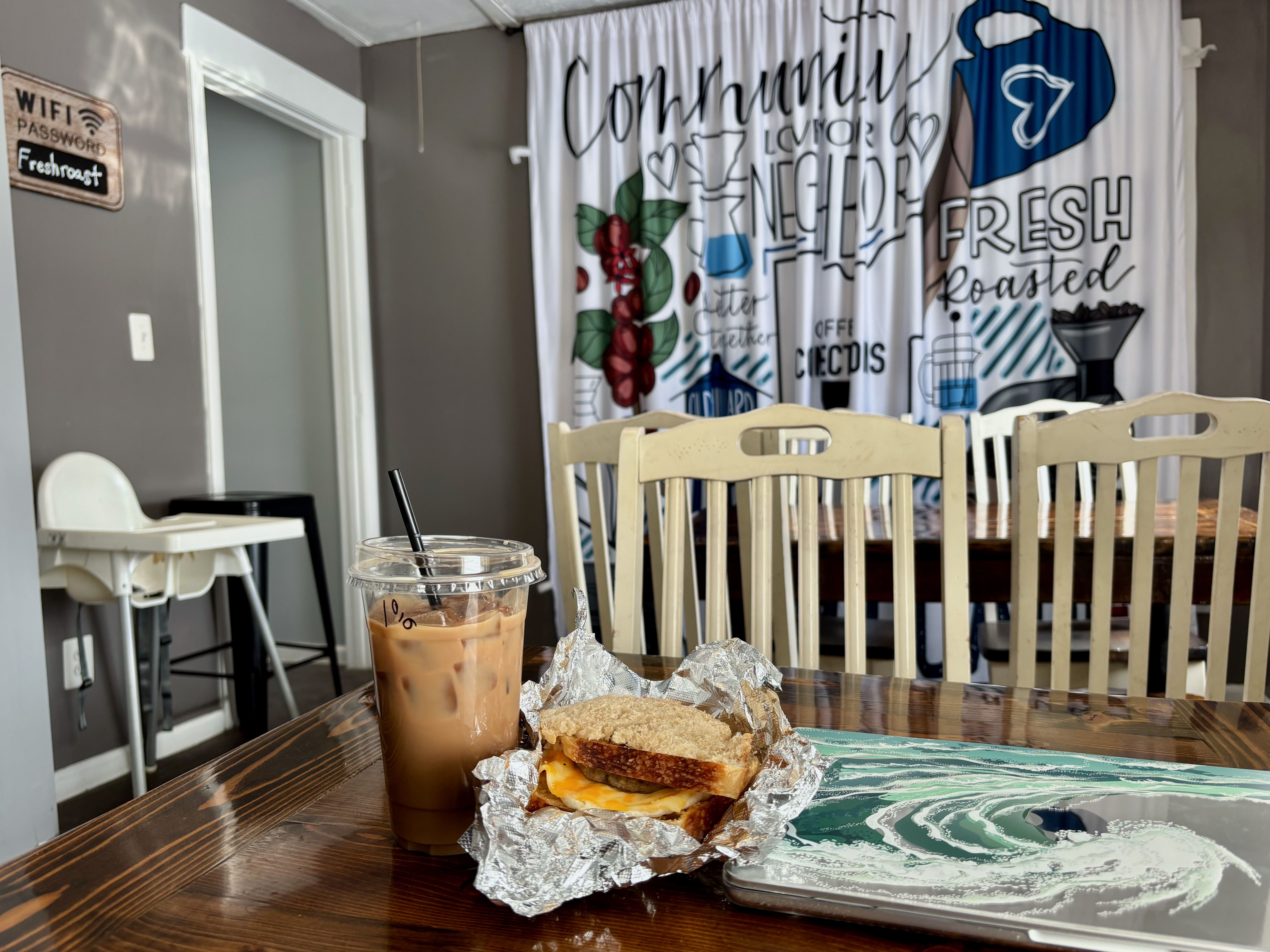A table with a laptop, iced coffee and breakfast sandwich in foil with a Coffee Connections-themed curtain in the background