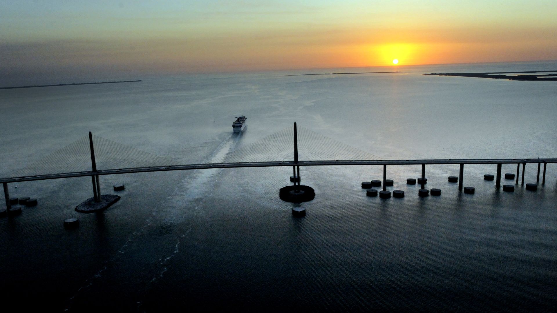 Sunset over calm water with a bridge stretching across it. A boat moves under the bridge, leaving a wake behind. Sky glows orange and blue near the horizon.
