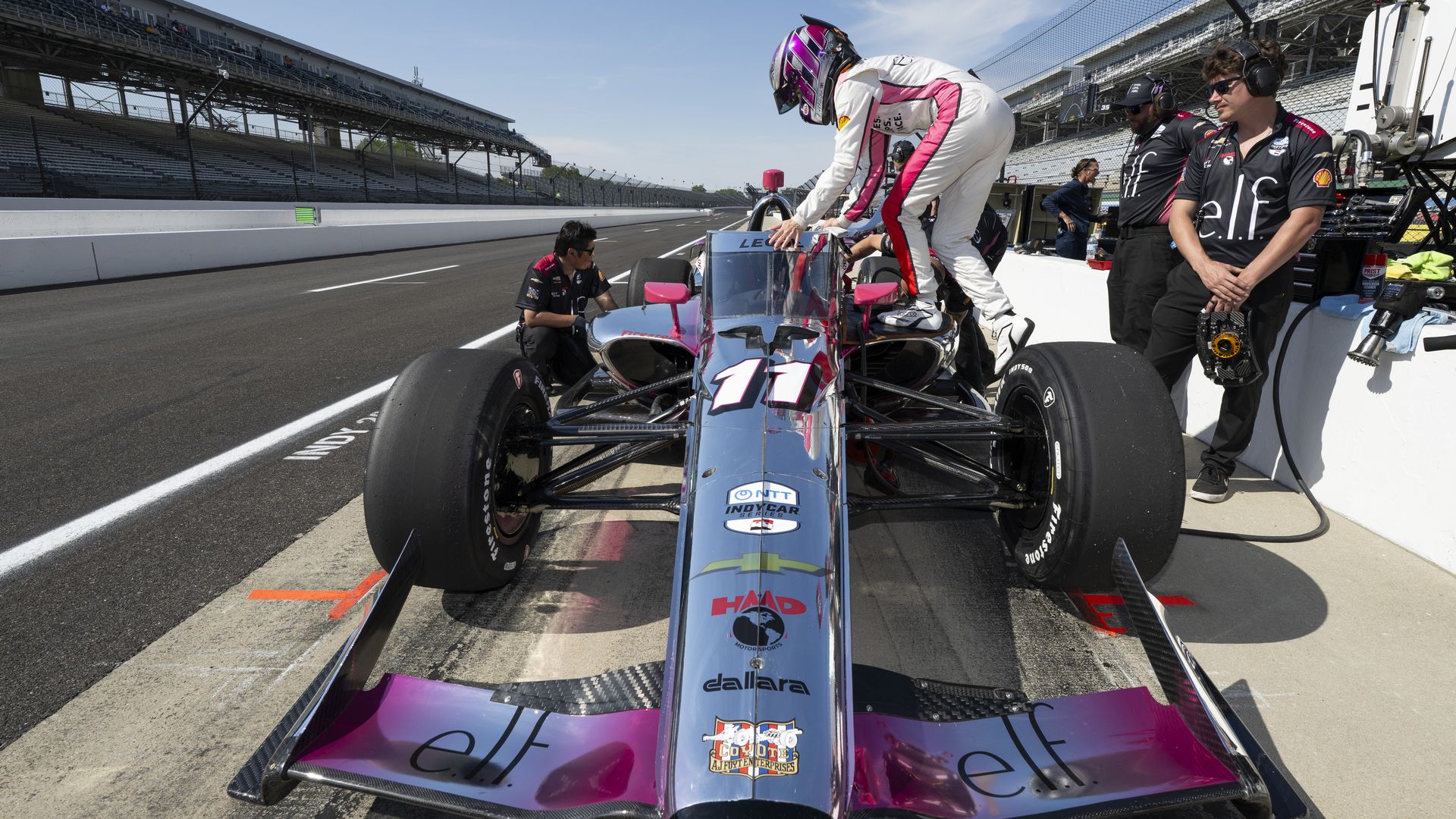 IndyCar in the pit lane with a driver in a white racing suit and pink helmet boarding car #11; crew in black ELF uniforms watch nearby as grandstands and blue sky loom in the background.