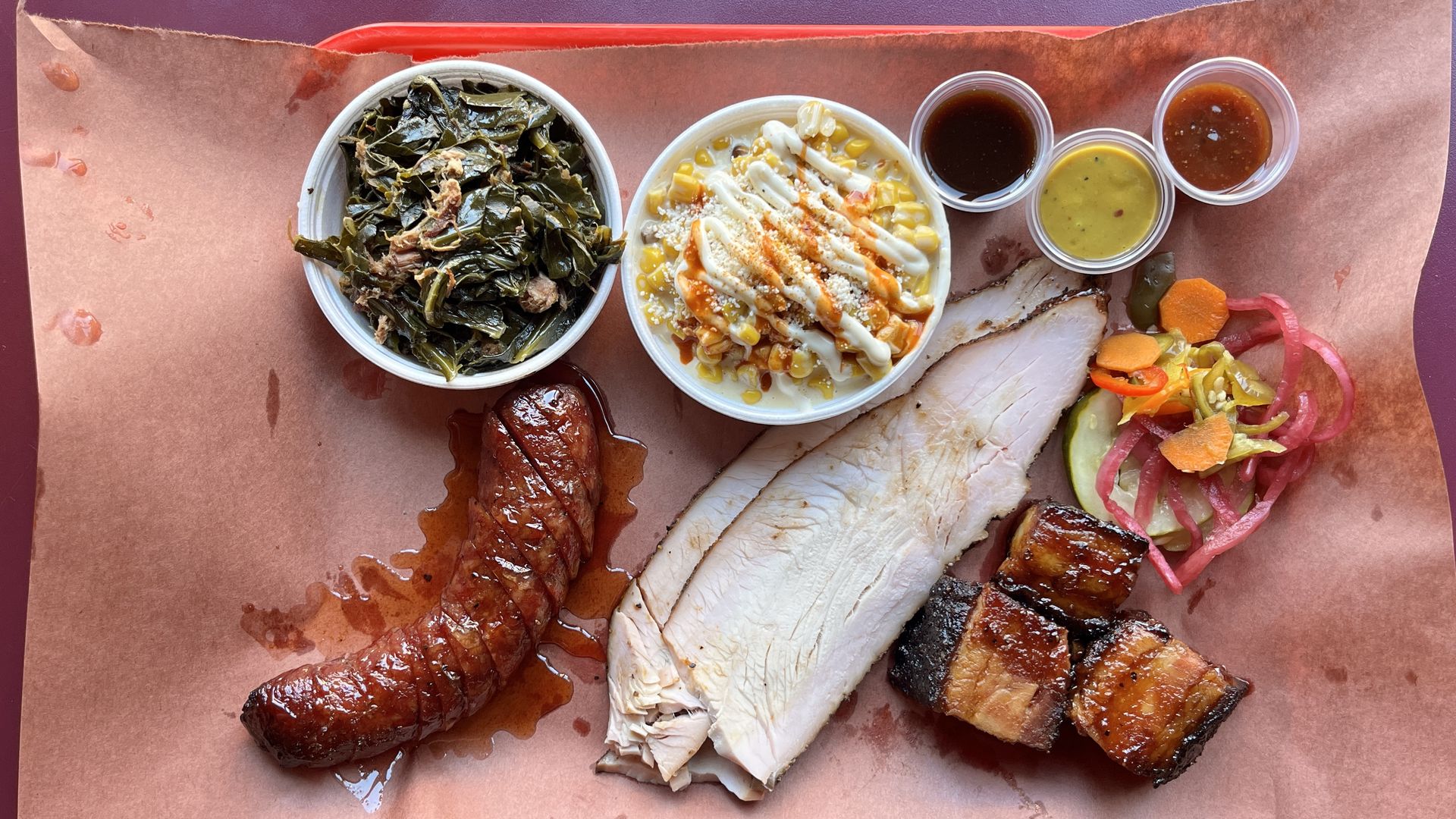 Barbecue platter with smoked sausage, sliced turkey, glazed bacon burnt ends, plus collared greens, elote corn, pickled vegetables, and three small cups of barbecue sauces on brown paper at Chef J.