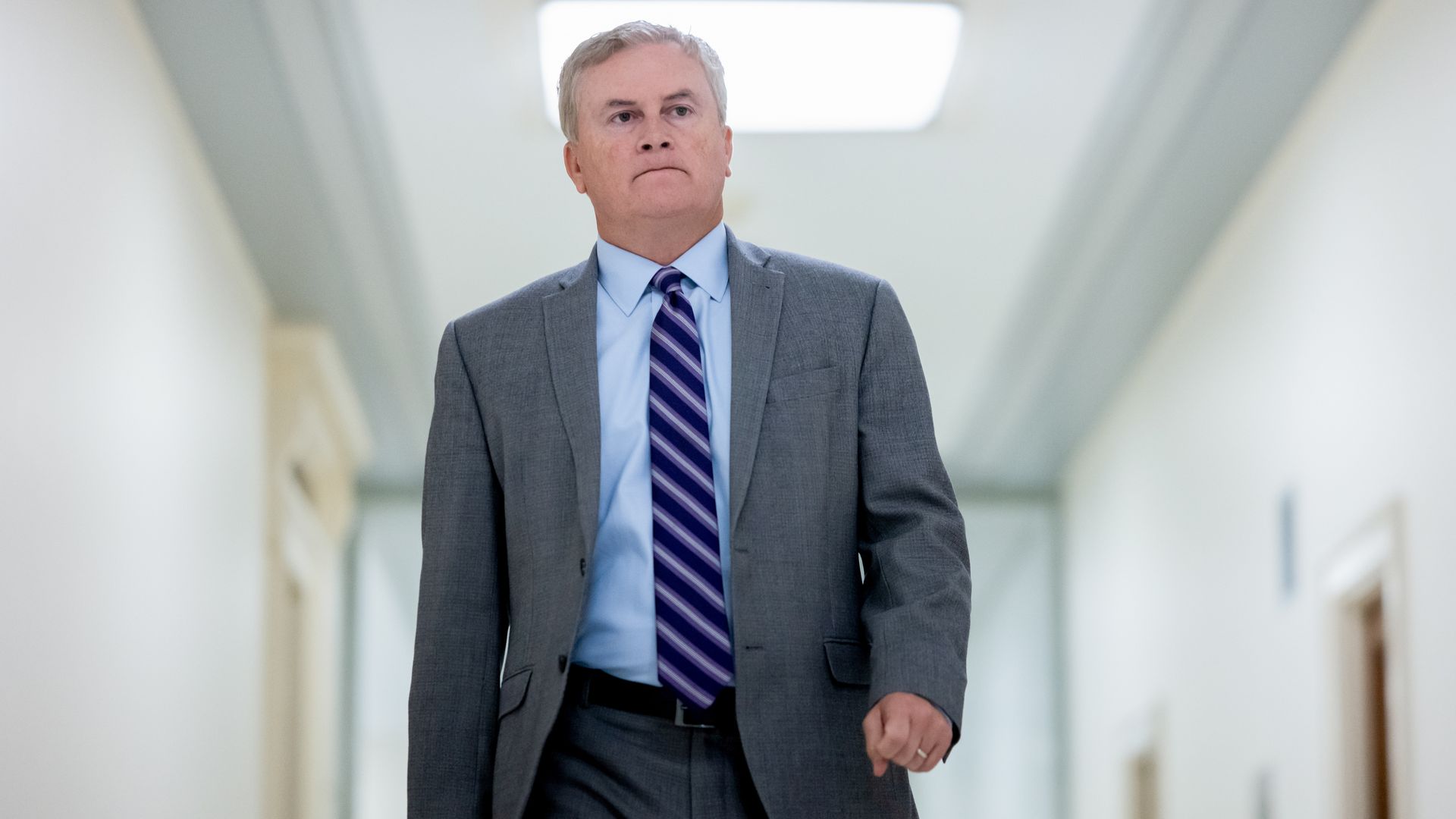 A middle-aged man in a gray suit, light blue shirt, and purple striped tie walks down a bright hallway, looking to the side.