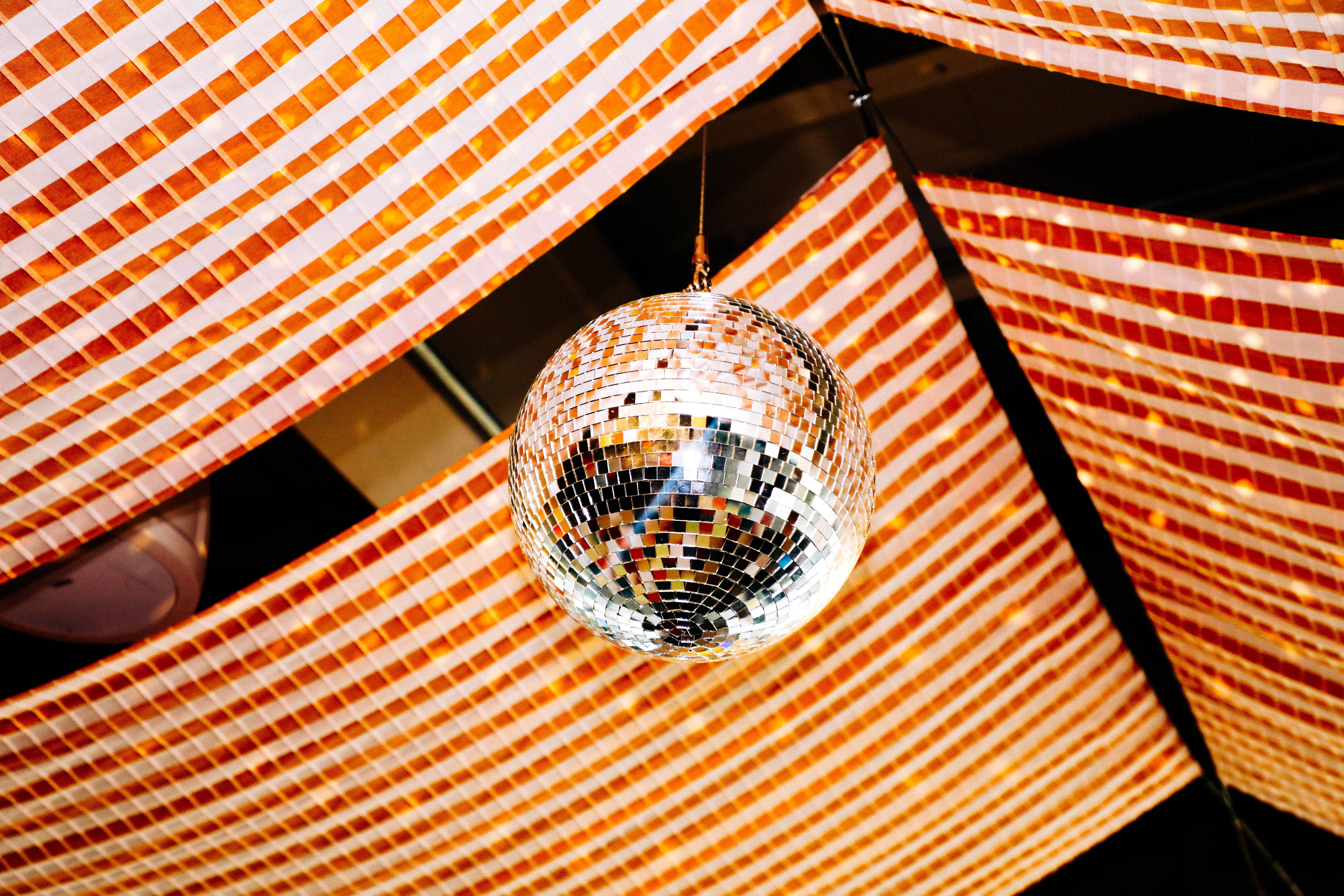A mirrored disco ball hangs from the ceiling, reflecting light, amid orange-and-white striped fabric banners arranged diagonally.