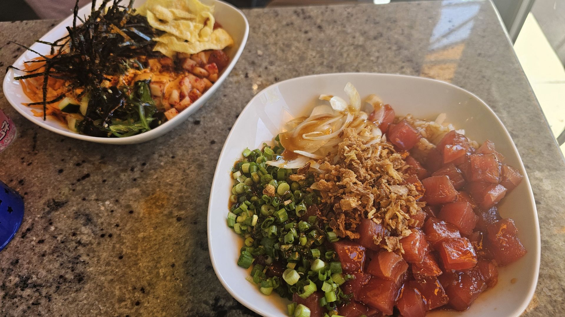 Two bowls of poke : one with diced raw fish, green onions, fried shallots, and onions; the other with seaweed, shredded carrots, cucumber, and wonton chips.