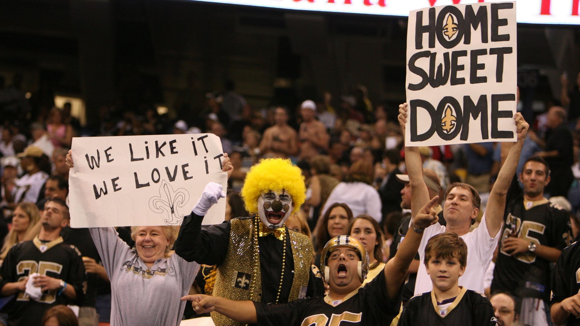 A photo of fans in the stands at the Superdome dressed in black and gold Saints gear. One holds a sign that says "Home Sweet Dome." Another has a sign that says "We like it, we love it."