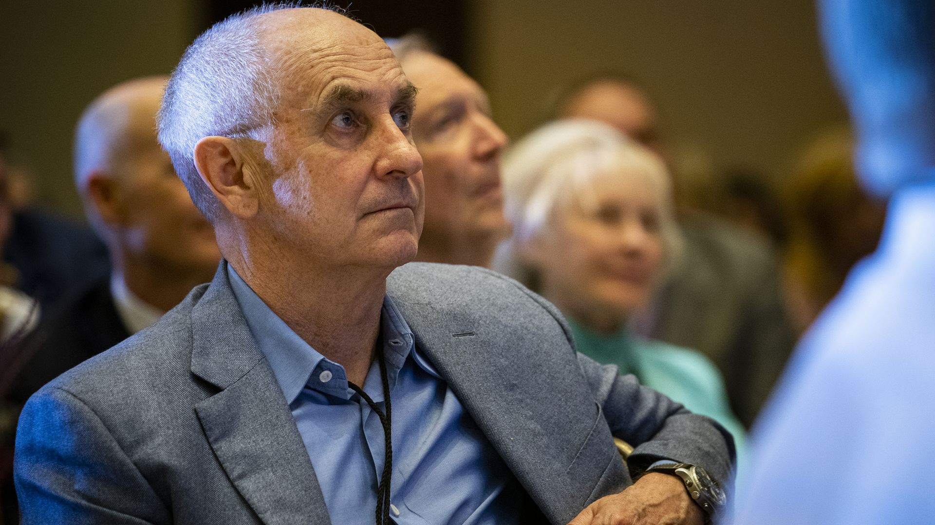 Person in a light blue shirt and gray blazer attentively listens at a formal event, with other adults blurred in the background.