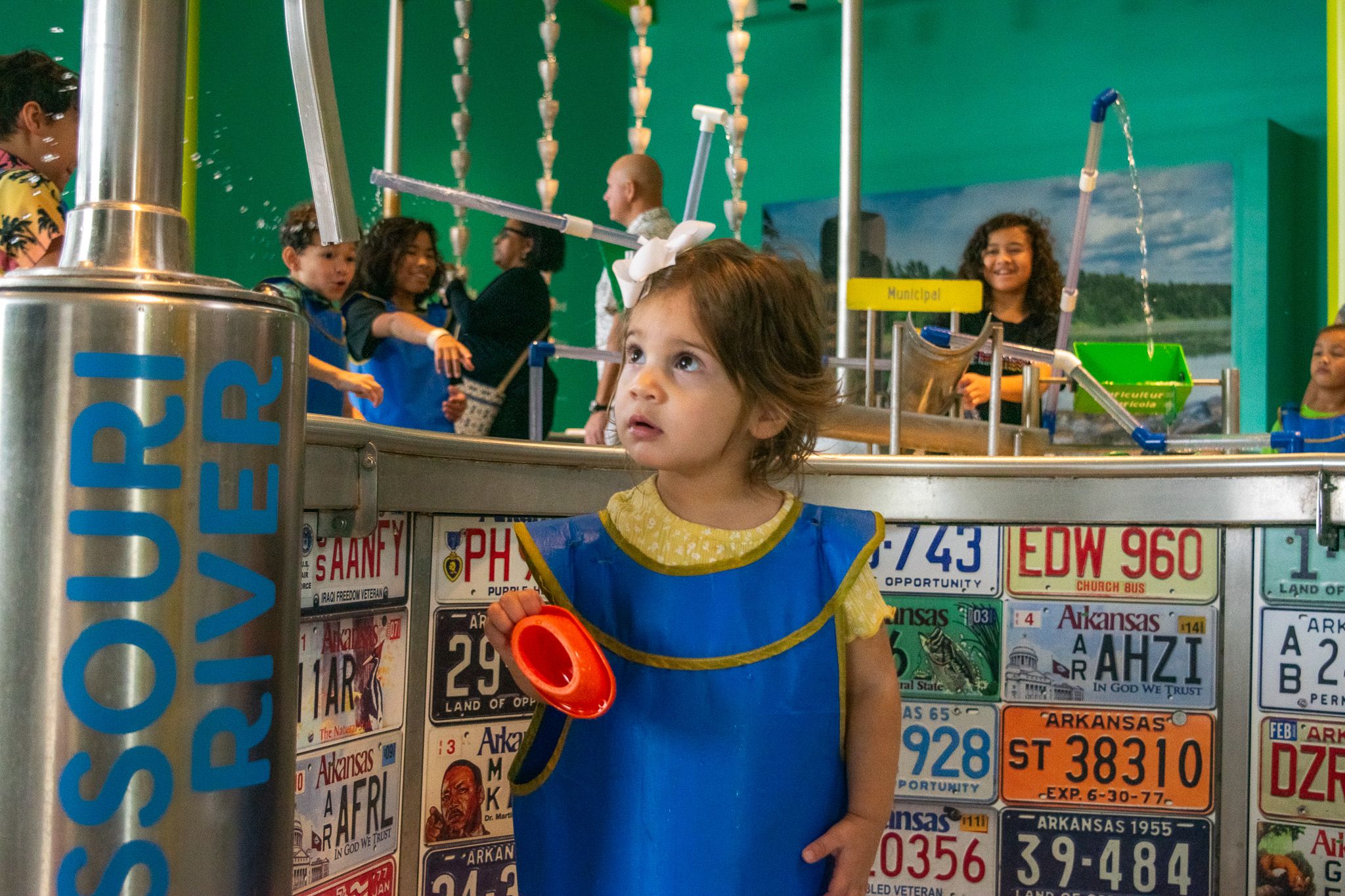 A young girl in a blue smock looks up at a water-park style exhibit with vertical pipes and flowing water, behind her a wall of colorful license plates and other kids playing.