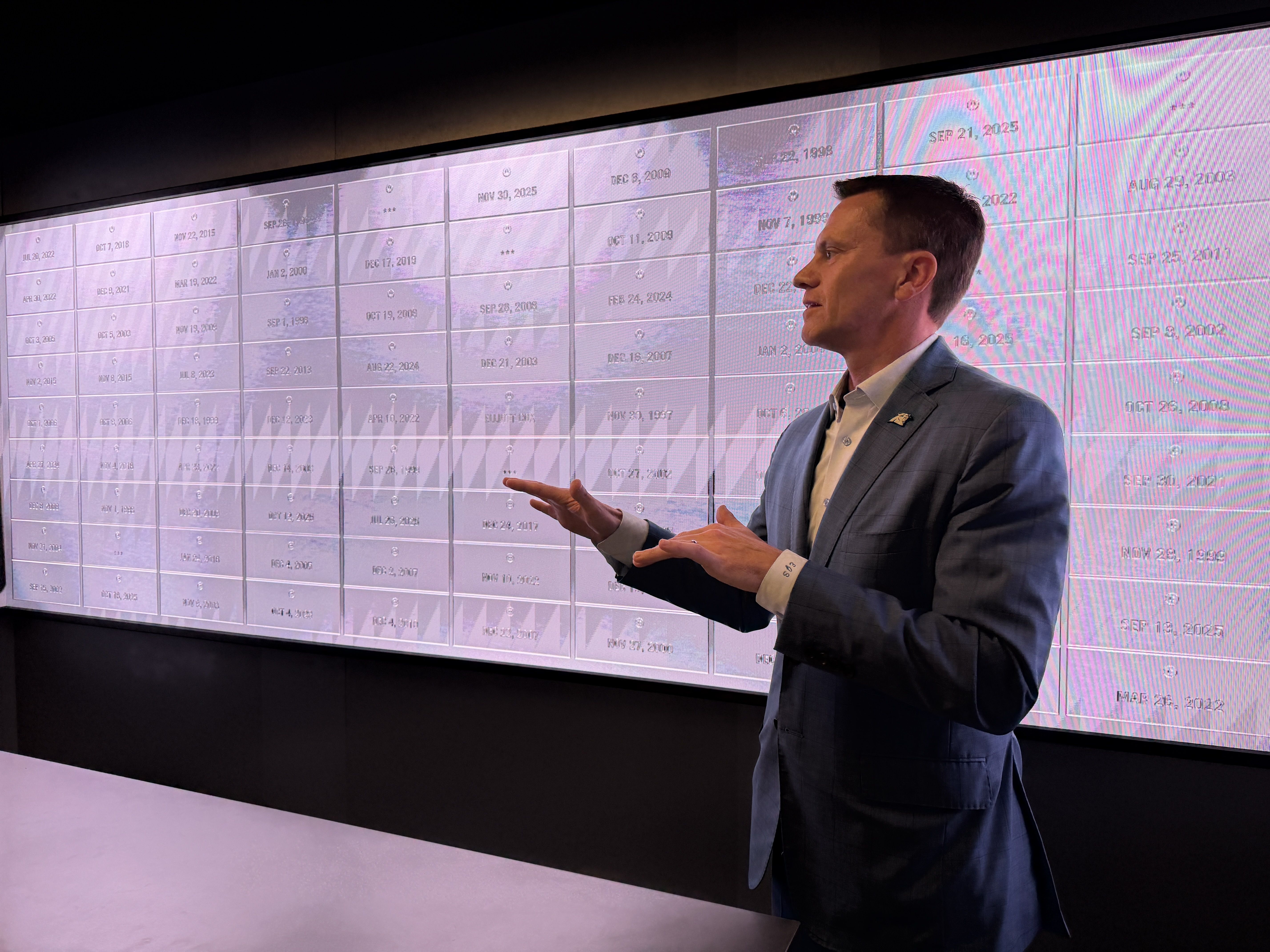 Side profile of a man in a light blue suit presenting beside a wall of calendar-style tiles showing dates; dark room, pink-tinted screen, gesturing with his hands.