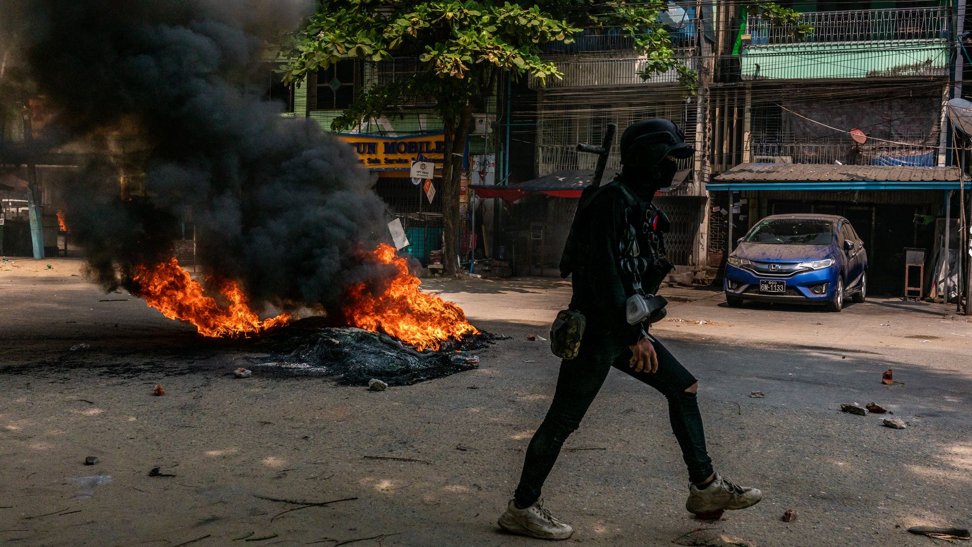 A person in military gear walks past a car on fire in the street.