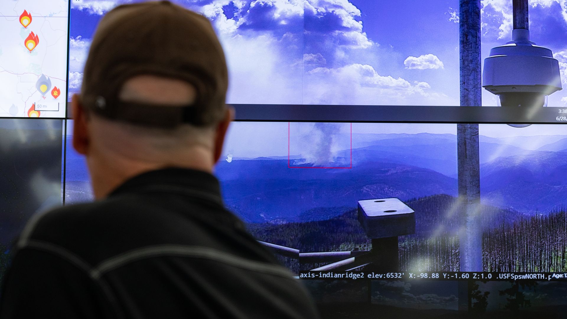 Back of a person with a cap watches a large wall of screens: a blue sky with mountains, a red-outlined area, a lamp post, and a map with flame icons; white data text runs across bottom.