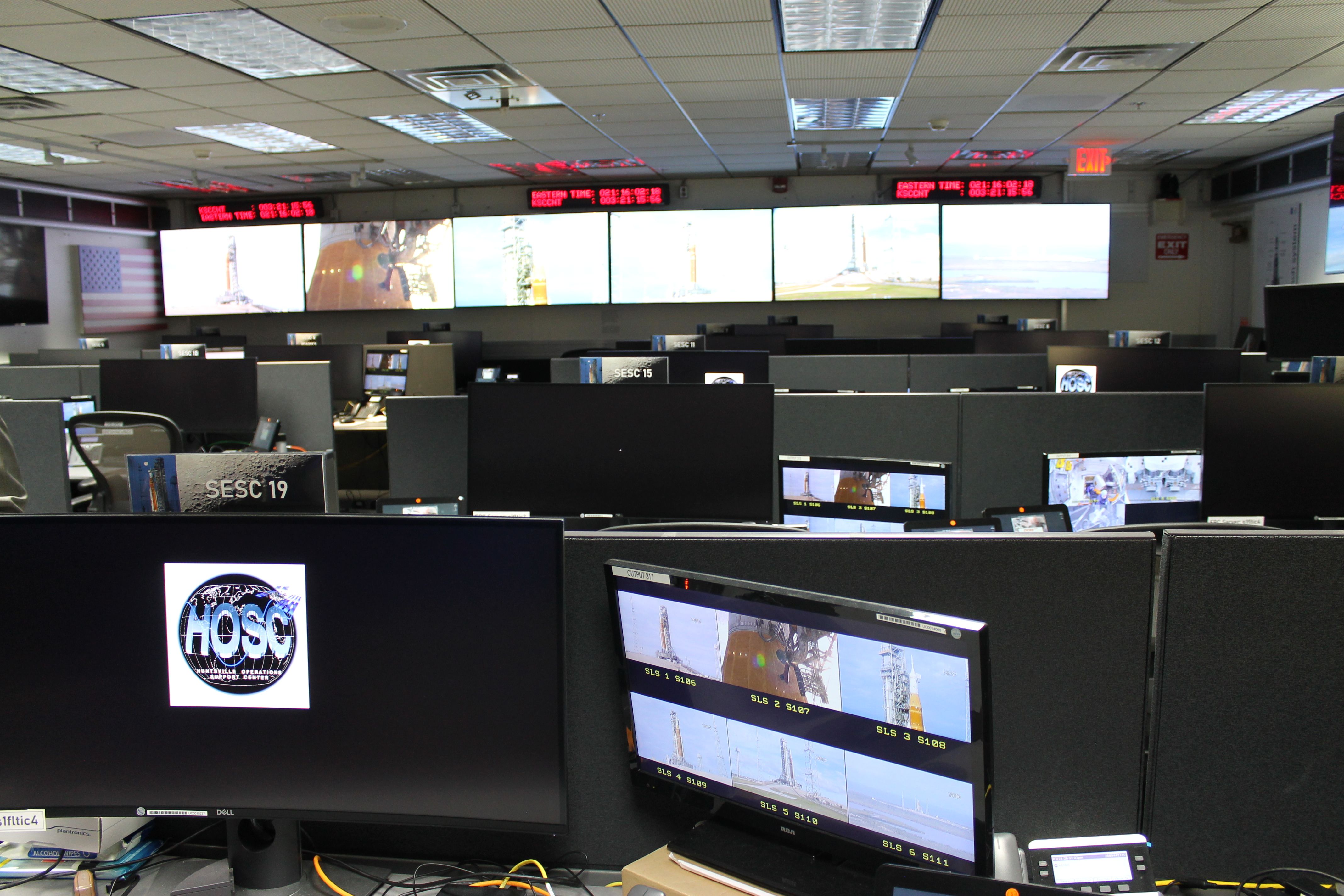 Room filled with computer monitors and cubicles displaying rocket launch images, with large screens on the far wall showing launch sequences and red digital clocks above them.
