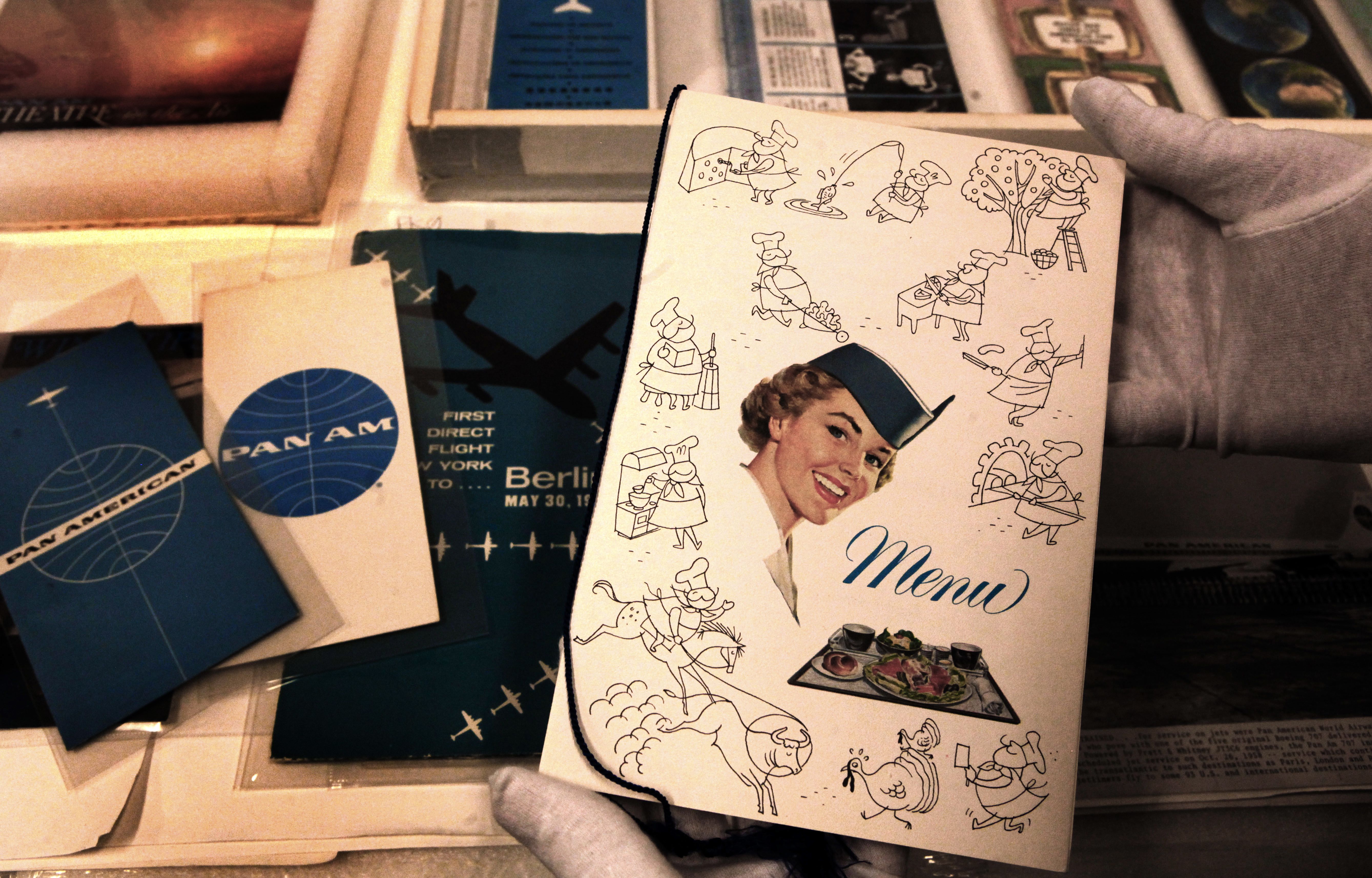 Vintage Pan Am menu held by gloved hands, featuring a smiling flight attendant, cartoon chefs, and a meal tray illustration, with blue and white Pan Am brochures in the background.