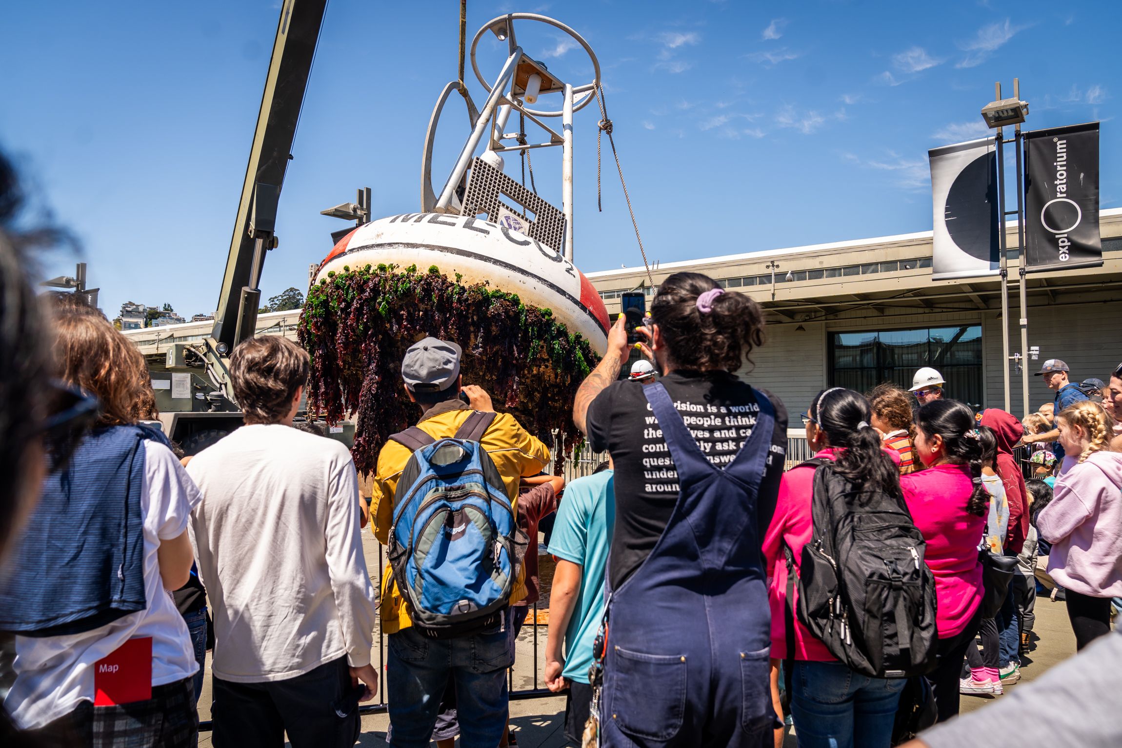 A group of people watch and photograph a large buoy covered in seaweed and marine growth being lifted by a crane outside a building with explorer-themed banners.