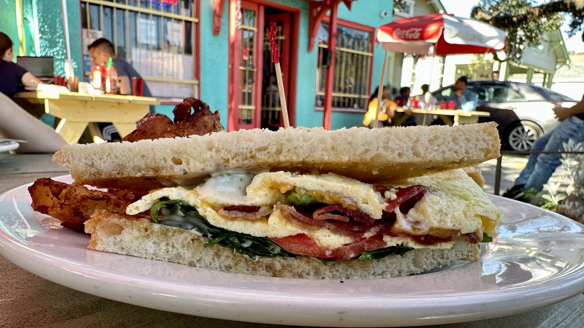 Photo shows a breakfast sandwich on a white plate with a turquoise restaurant in the background.