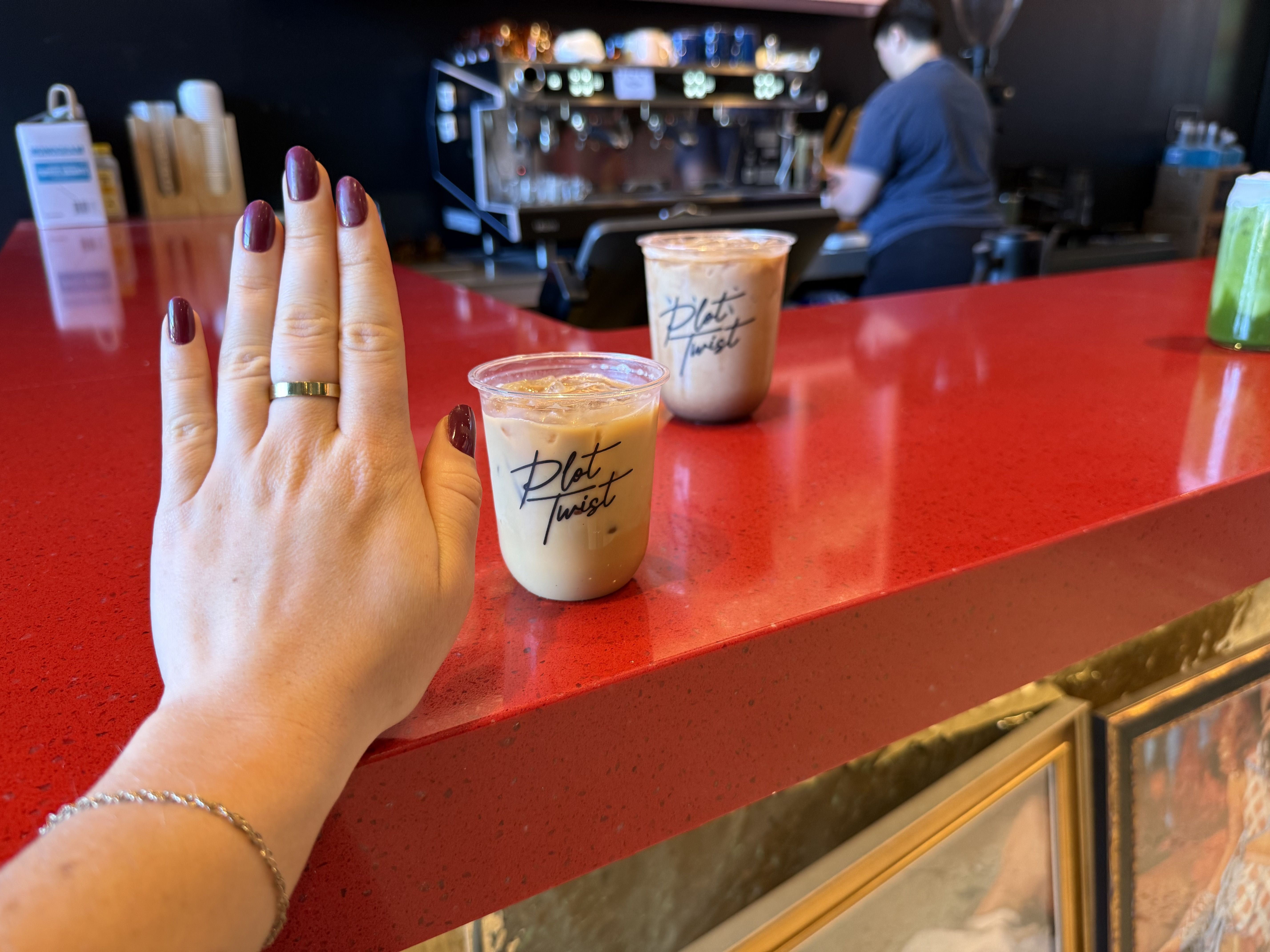 Close-up of a hand with burgundy nails and a gold ring resting on a bright red counter. Two iced coffees labeled "Plot Twist" with a barista and an espresso machine blurred in the background.