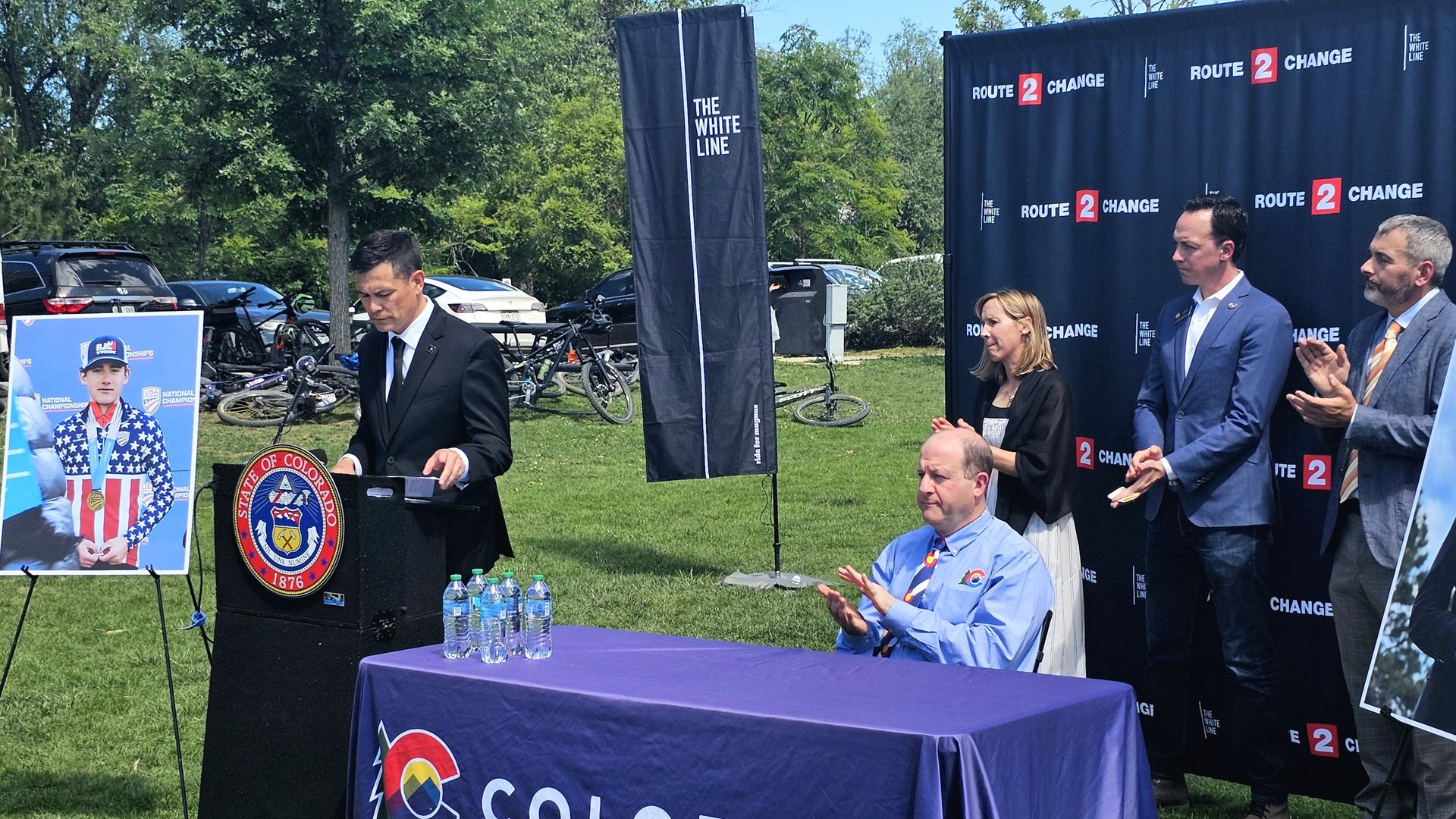 Outdoor press event with speaker at podium bearing the Colorado seal; backdrop reads Route 2 Change; suited officials stand nearby; left poster features a patriotic cyclist; water bottles on a table.