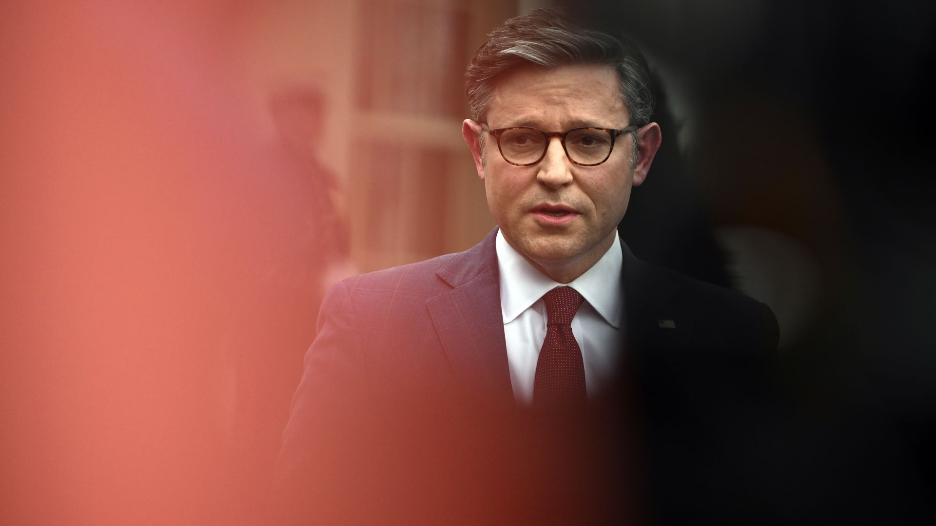 House Speaker Mike Johnson, wearing a dark blue suit, white shirt and red tie, in a lens flare.
