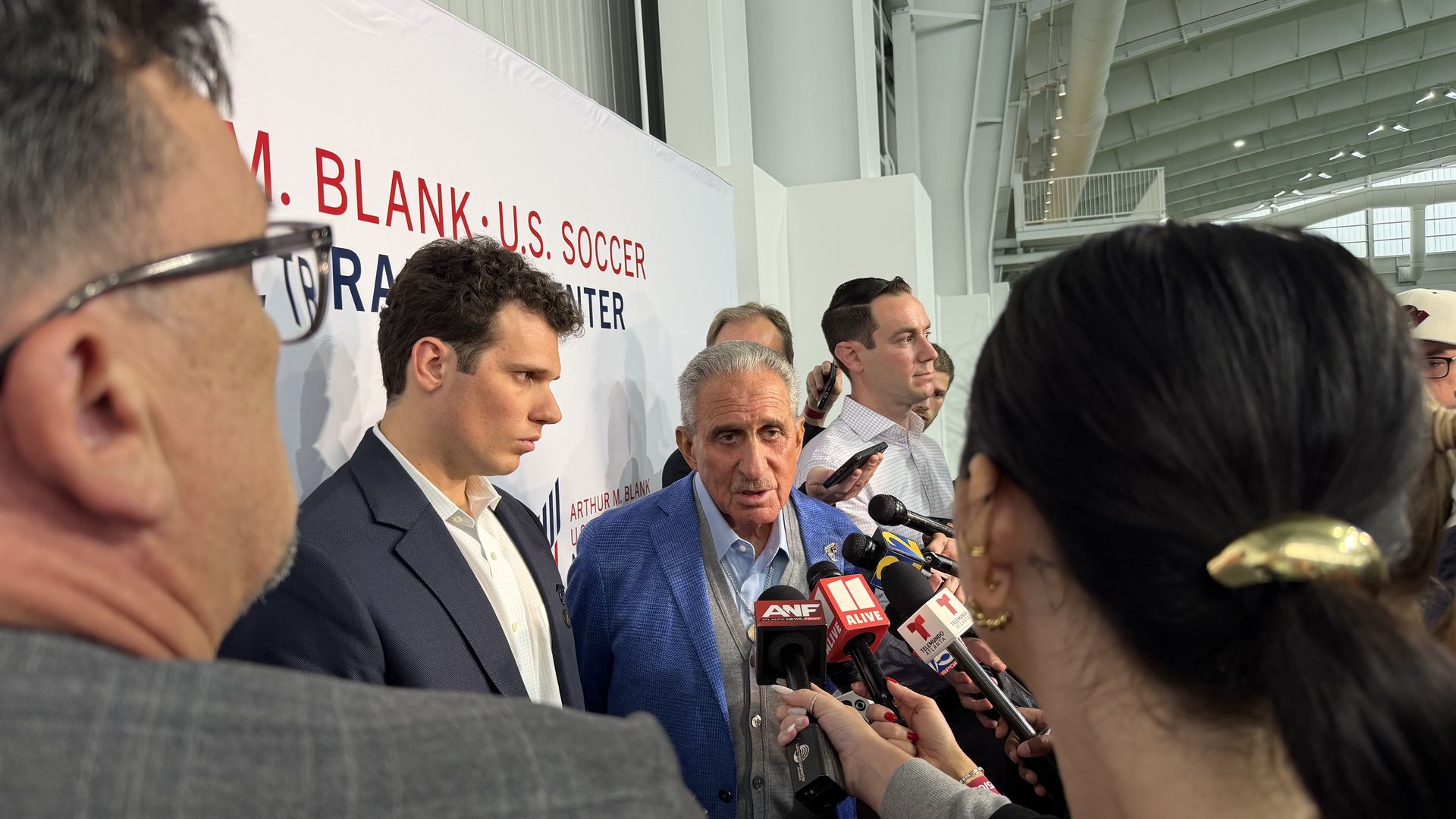 A press conference: a man in a blue blazer speaks to reporters holding microphones; a large banner reads "U.S. Soccer" behind him, amid a crowd and glass-structured arena.
