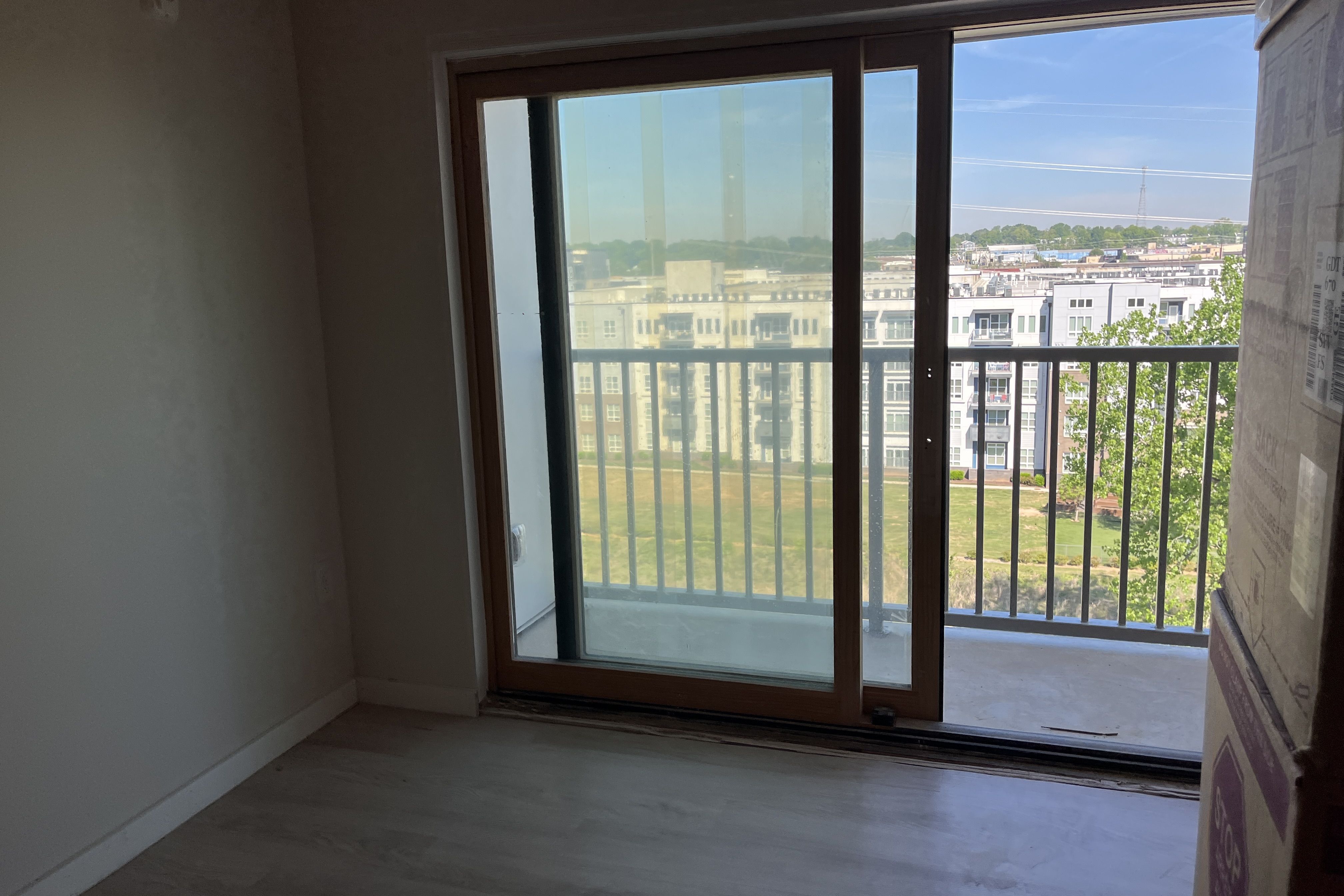Empty room with a large sliding glass door opening to a balcony. Brown wooden frame, light floor, and bare walls. Outside, city buildings, green trees, and a blue sky with power lines; a cardboard packaging panel sits to the right.