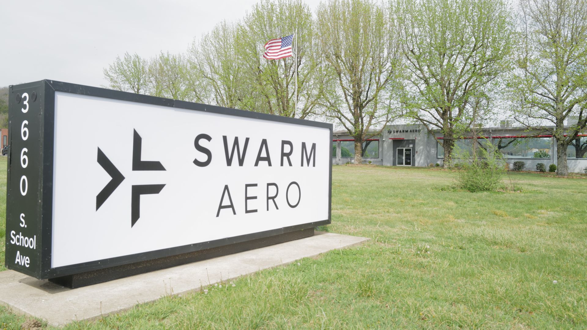 Wide outdoor scene with a large white sign reading 'SWARM AERO' and a black arrow. In the background are an American flag, green trees, and a gray building.