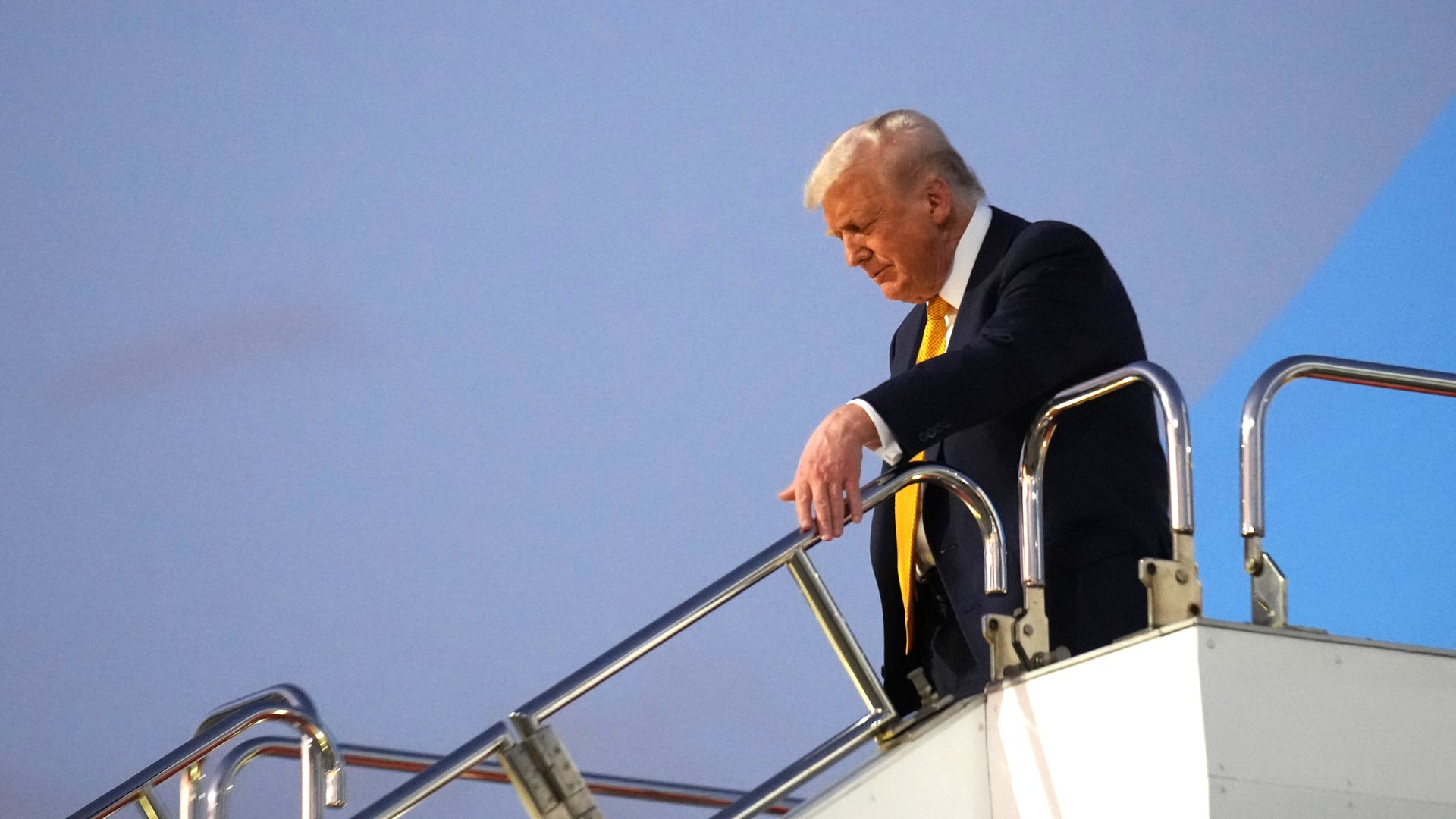 President Trump — wearing a yellow tie, a white collared shirt and a dark suit — holds onto the hand rail as he descends the stairs from Air Force One.