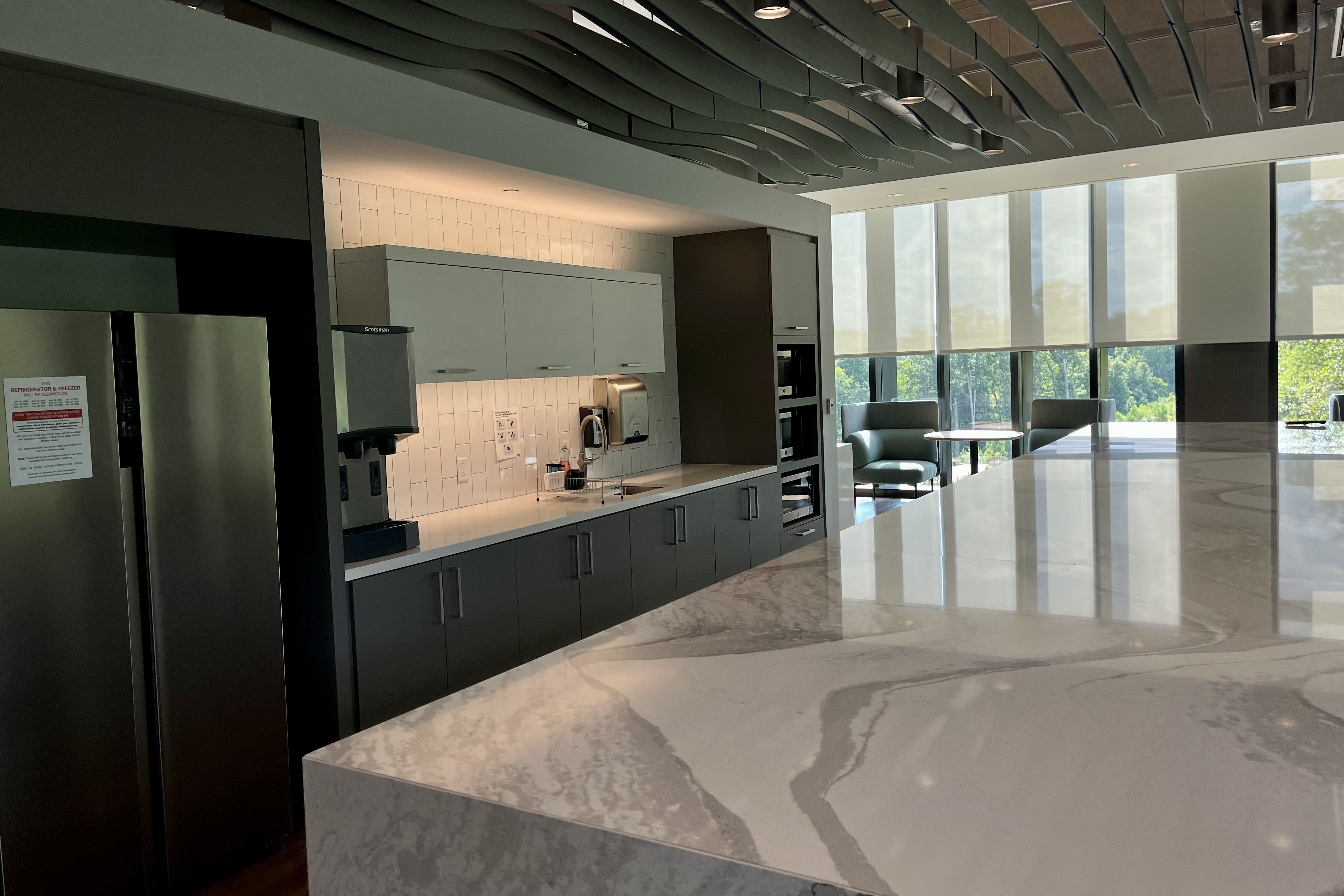Modern kitchen area with gray and white cabinetry, stainless steel refrigerator, large marble countertop, sink, coffee machine, and seating area with green chairs by large windows.