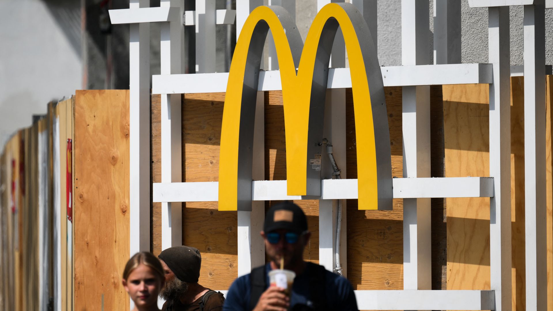 Bright yellow McDonald's arches rise behind white slats and plywood fencing at a construction site; in the foreground a person with a drink and a person wearing sunglasses walk by.