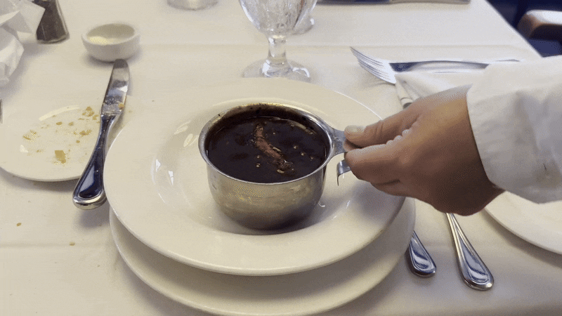 A hand pours a silver container of gumbo into a white bowl and retreats.