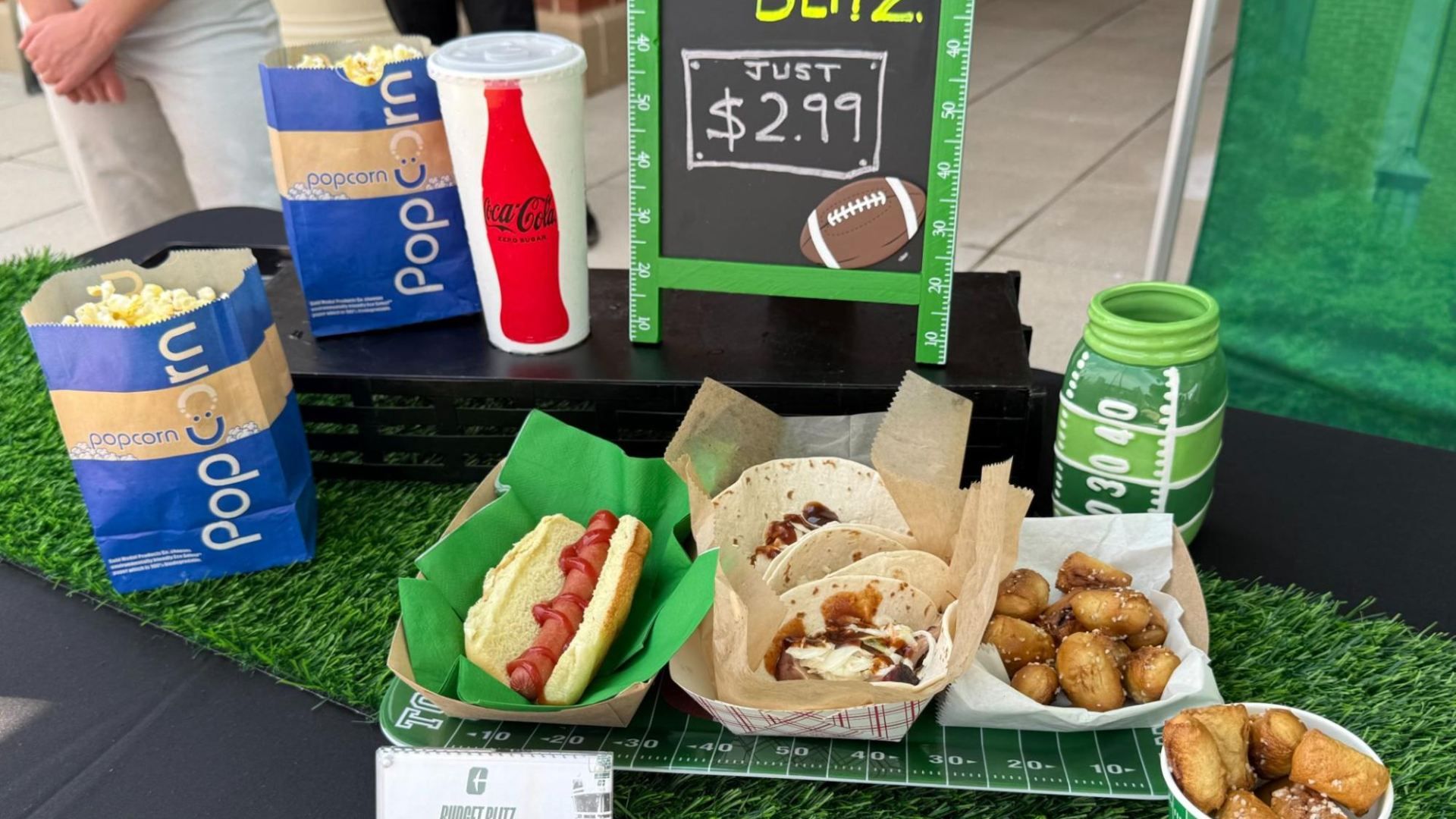 Snack setup on artificial turf with blue popcorn bags, a Coca-Cola can, hot dogs, tacos, pretzels, and a football-themed sign showing "Just $2.99."