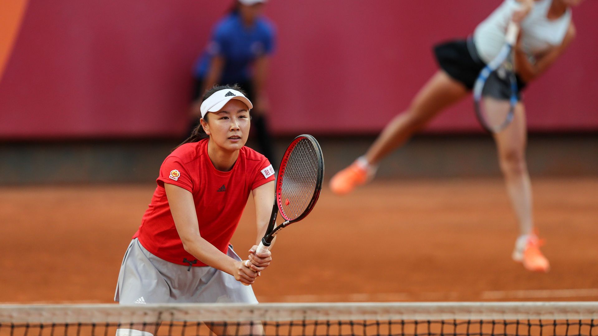 Photo of Peng Shuai crouched with her racket in front of her on a tennis court as her partner serves from behind