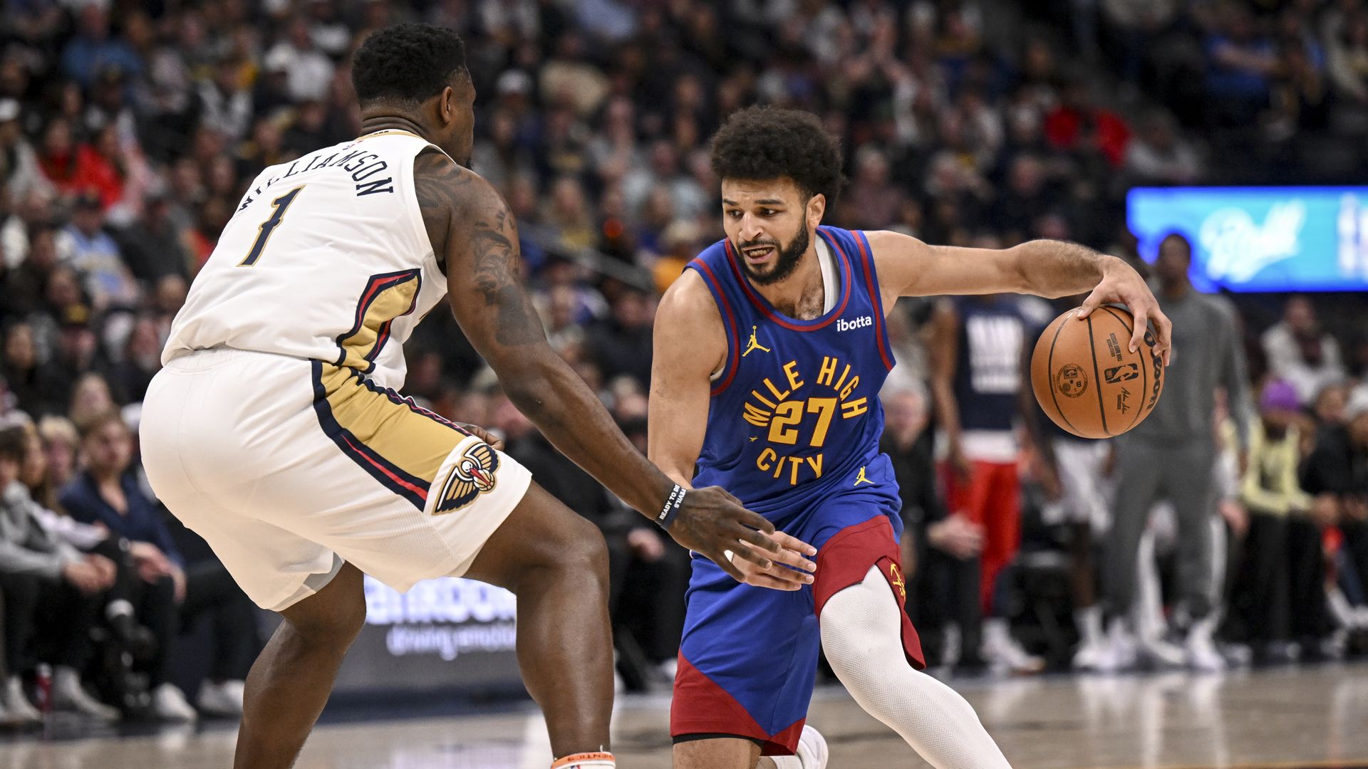 A man dribbles a basketball in a blue uniform while another man in white uniform stands in front of him.