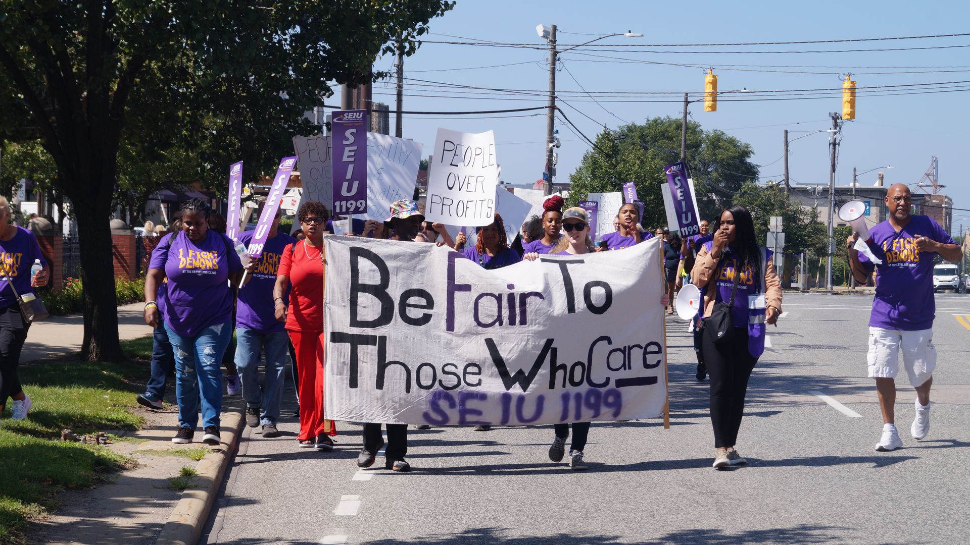 Diverse protesters march on a sunny street, holding a large banner reading "Be Fair To Those Who Care" with signs like "People Over Profits." Purple SEIU 1199 banners and a megaphone are visible.