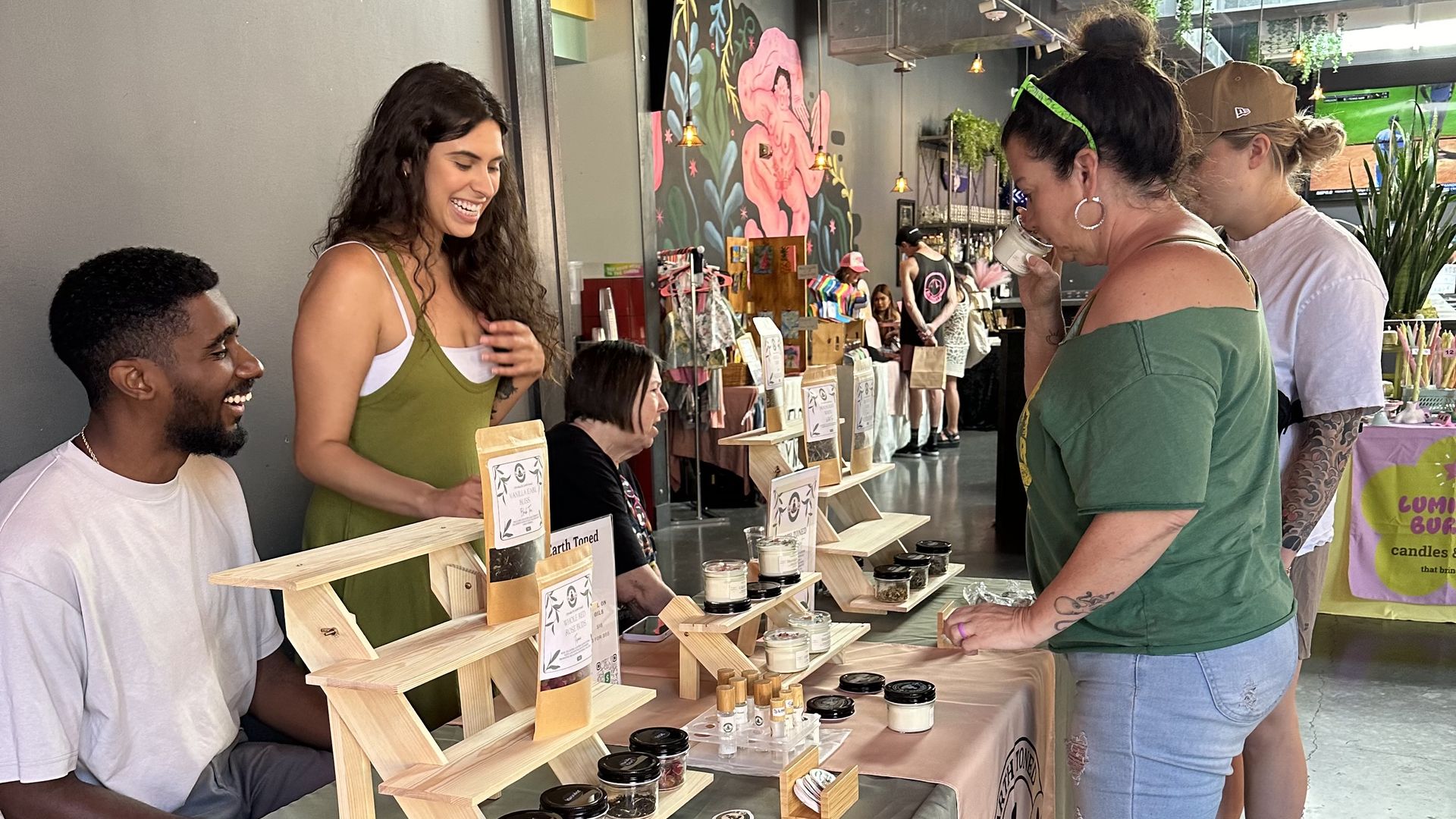 Two vendors seated behind a table with candles and stickers on display, smiling at two women customers, one of whom is smelling a candle in a bright indoor market with floral wall art.