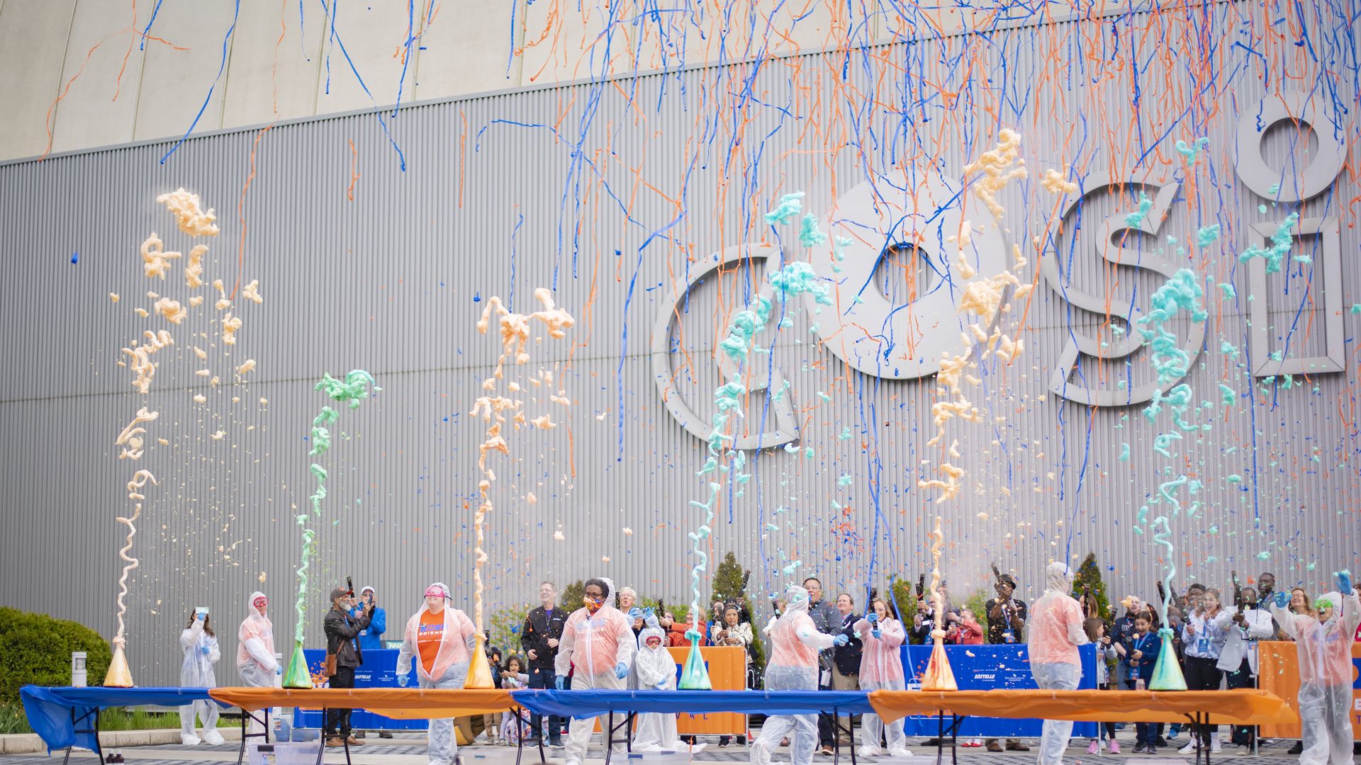 Outdoor celebration with orange, blue, and teal streamers and confetti; crowd in ponchos stands behind an orange‑covered table on steps, in front of a gray ribbed wall.