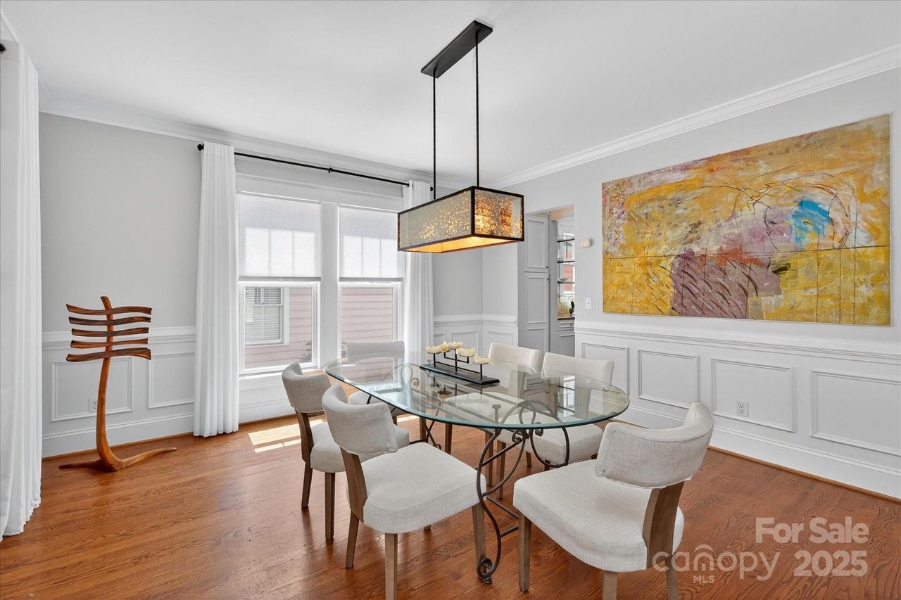 Bright dining room with glass oval table, six beige upholstered chairs, wooden floor, modern rectangular pendant light, white walls with paneling, large colorful abstract painting, and windows with white curtains.