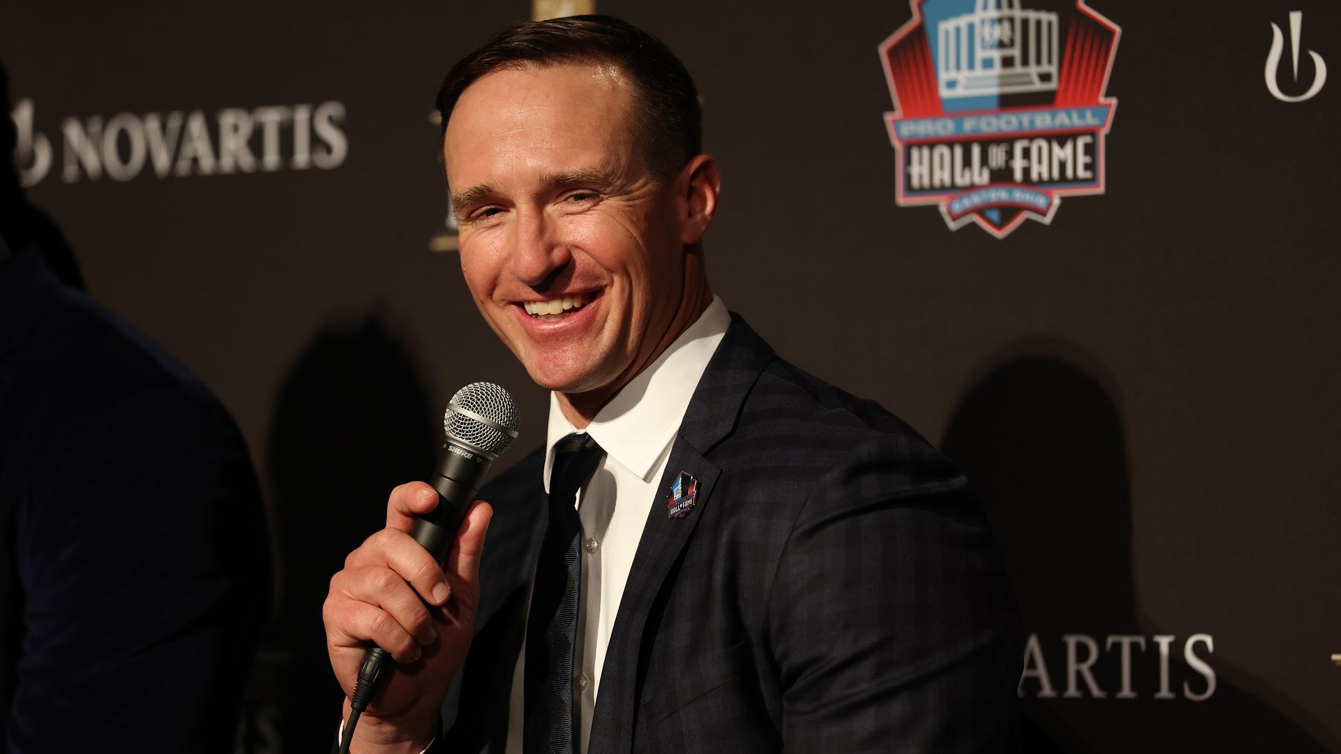 Image shows Drew Brees smiling. He's wearing a suit and holding a microphone.