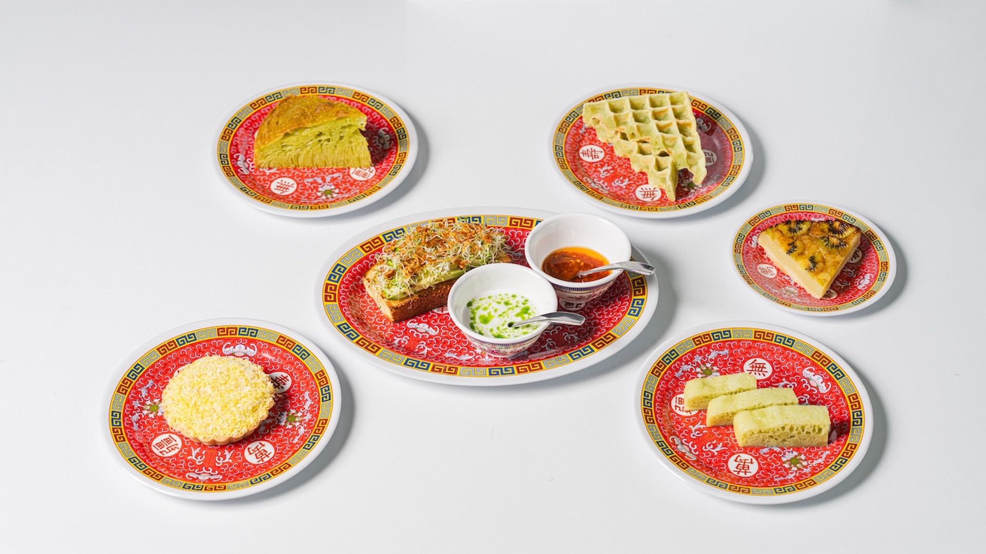 A colorful spread of dishes on traditional Chinese-patterned plates featuring various pastries, waffles, avocado toast topped with sprouts, and small bowls of sauces on a bright white surface.
